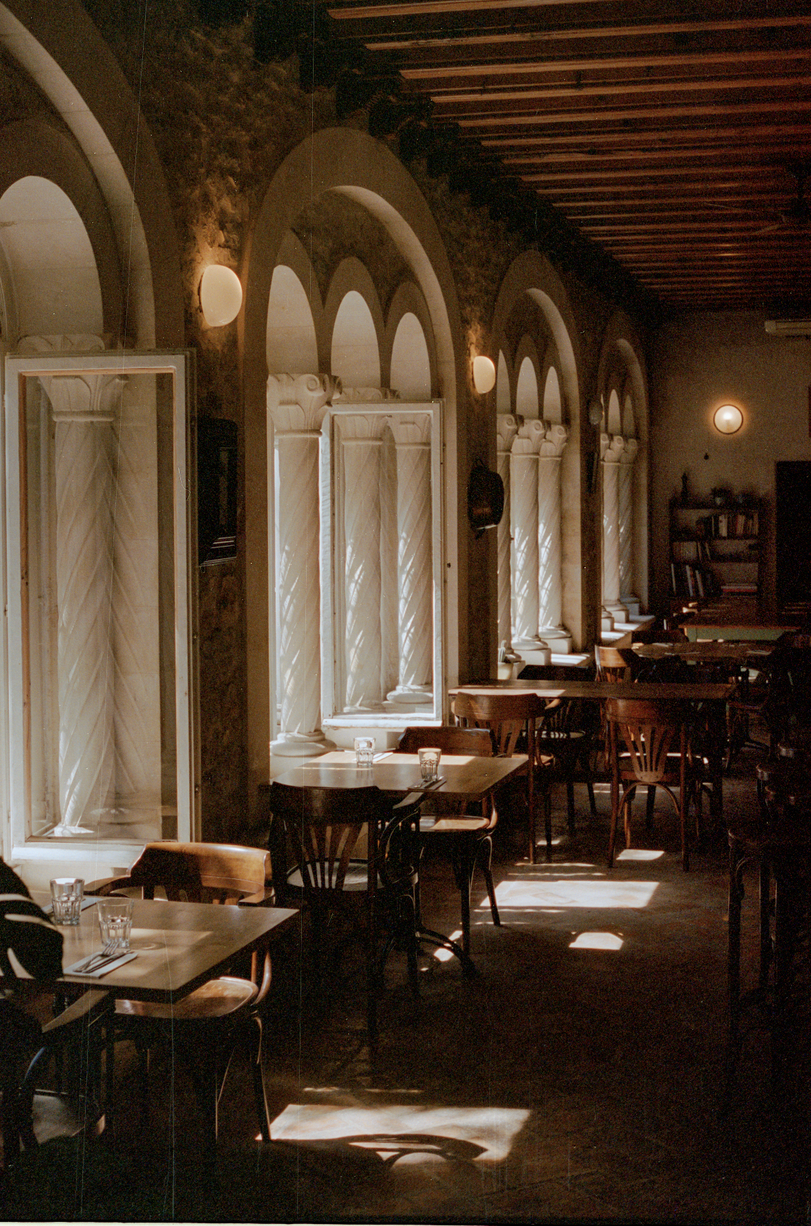 Interior of a dimly lit restaurant with wooden tables and chairs, large windows with white curtains, and elegant arched window frames along the wall. Sunlight filters through the curtains, creating patterns on the floor.