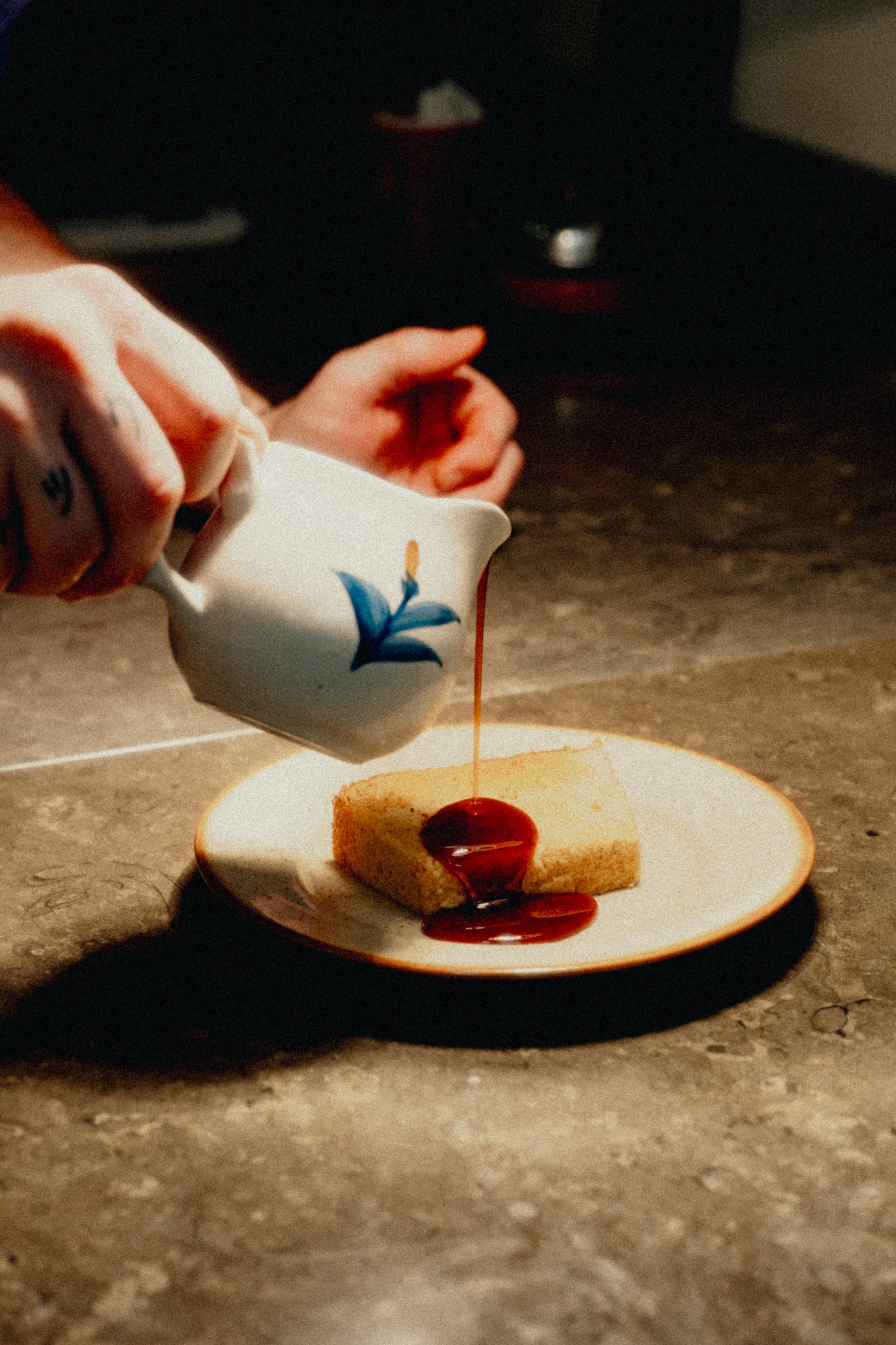 A hand pours red sauce from a small white teapot with blue floral design onto a slice of sponge cake on a white plate.