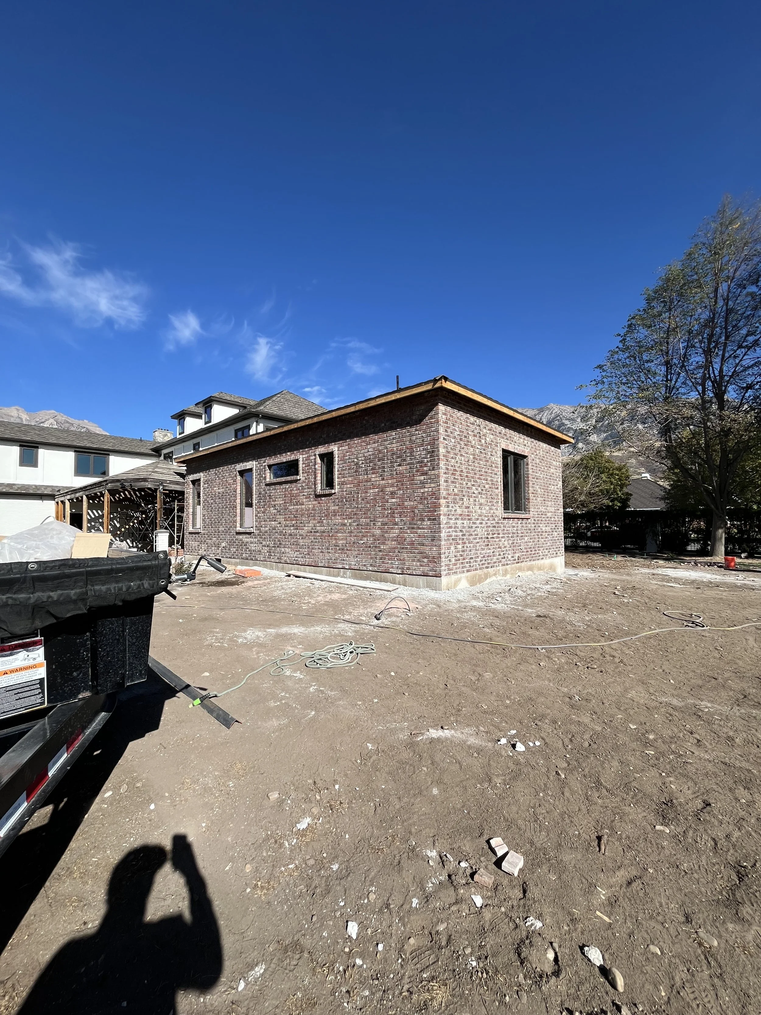 A brick house under construction with a clear blue sky, barren yard, construction equipment, and a shadow of a person taking the photo.
