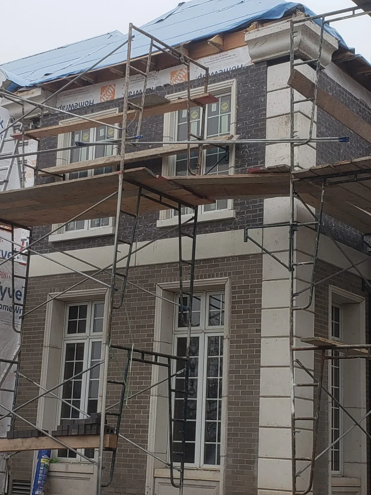 Construction scaffolding erected around a multi-story brick building under renovation, with windows visible and a blue tarp covering the roof.