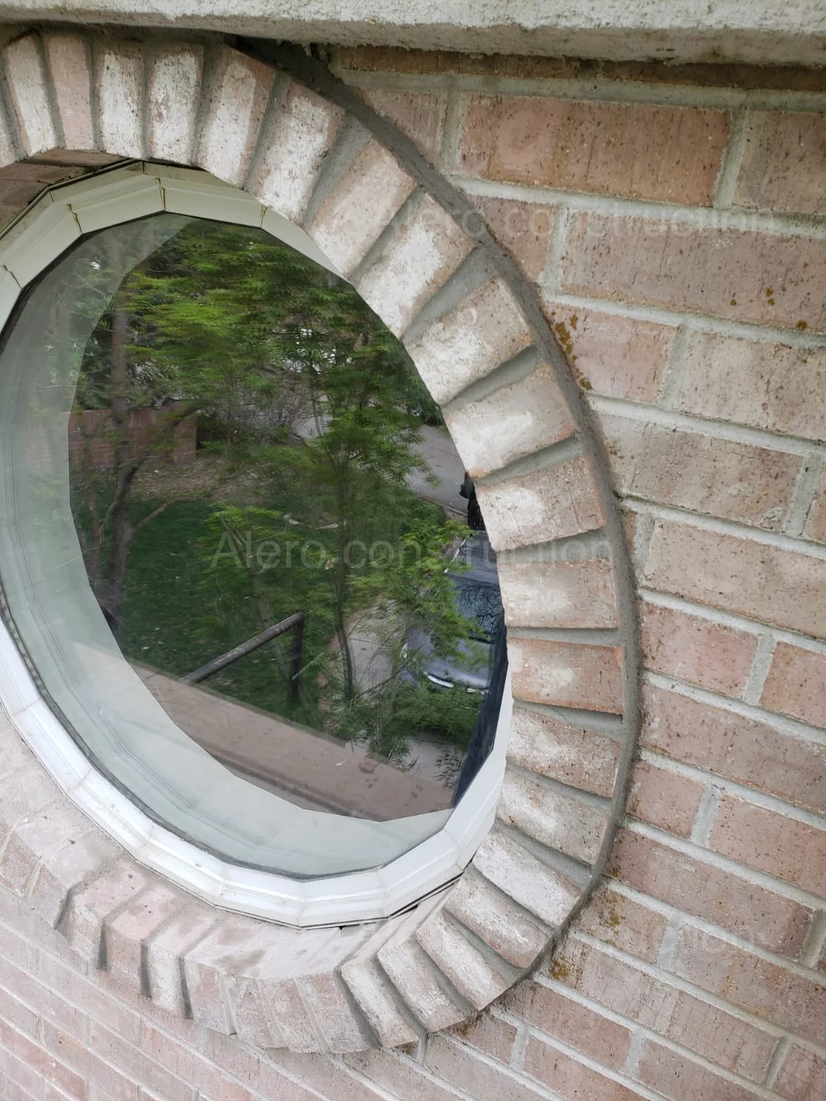 Rounded brick window with a reflective glass pane, showing a green leafy tree and parked car outside.