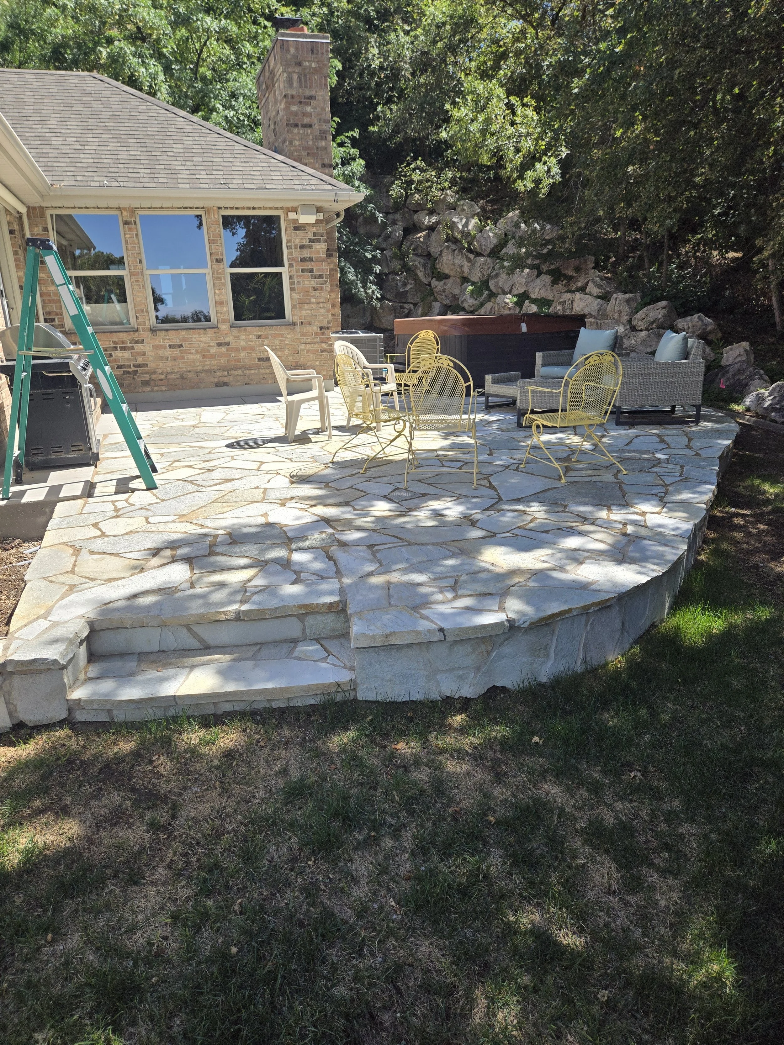 A backyard patio with a stone surface, outdoor furniture including yellow and gray chairs and a sofa, a hot tub, a small grill, and a ladder, with a brick house wall and trees in the background.