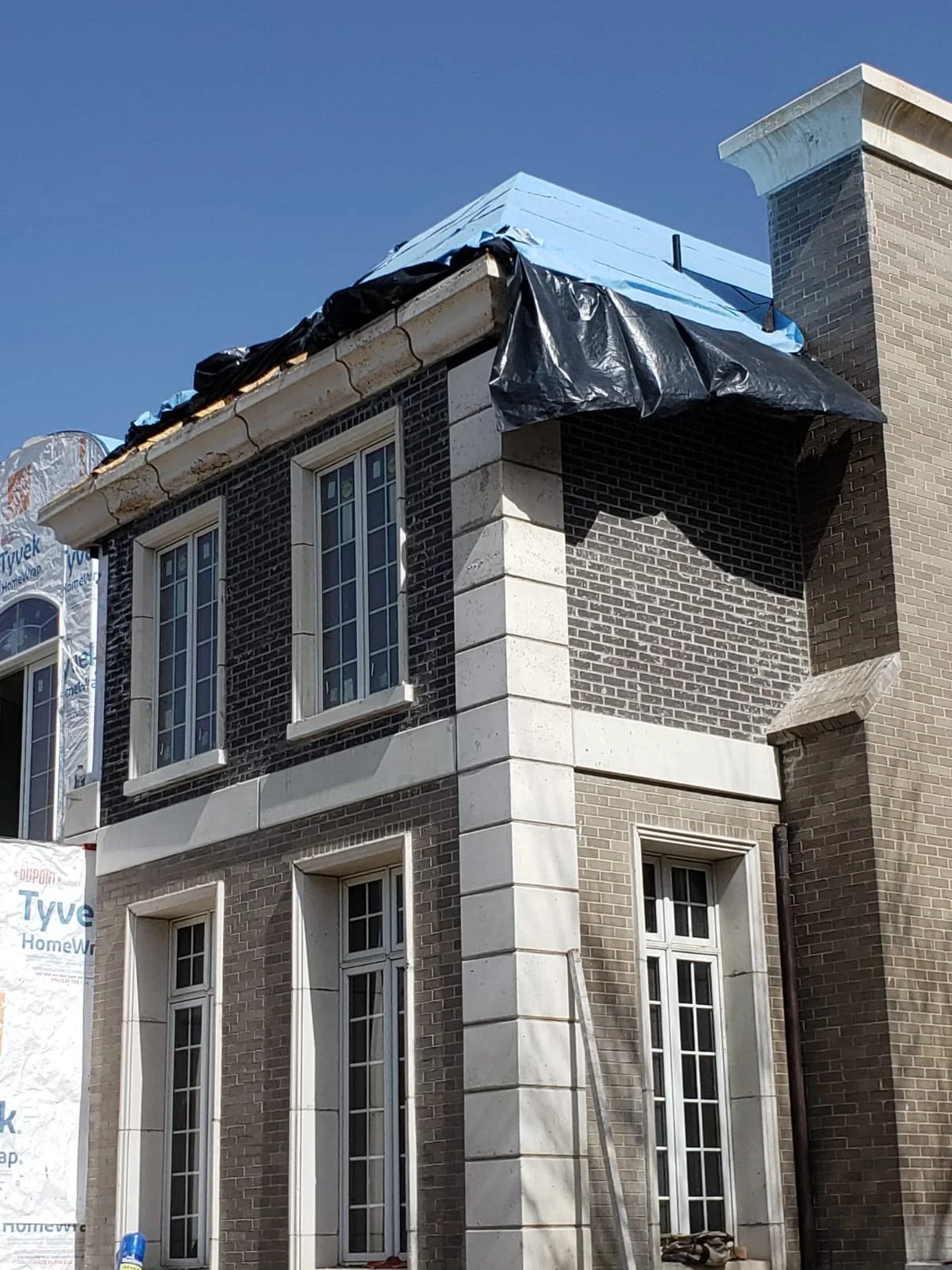 A two-story brick house under construction, with black plastic tarp and blue cover on the roof, white window frames, and a partially finished exterior.