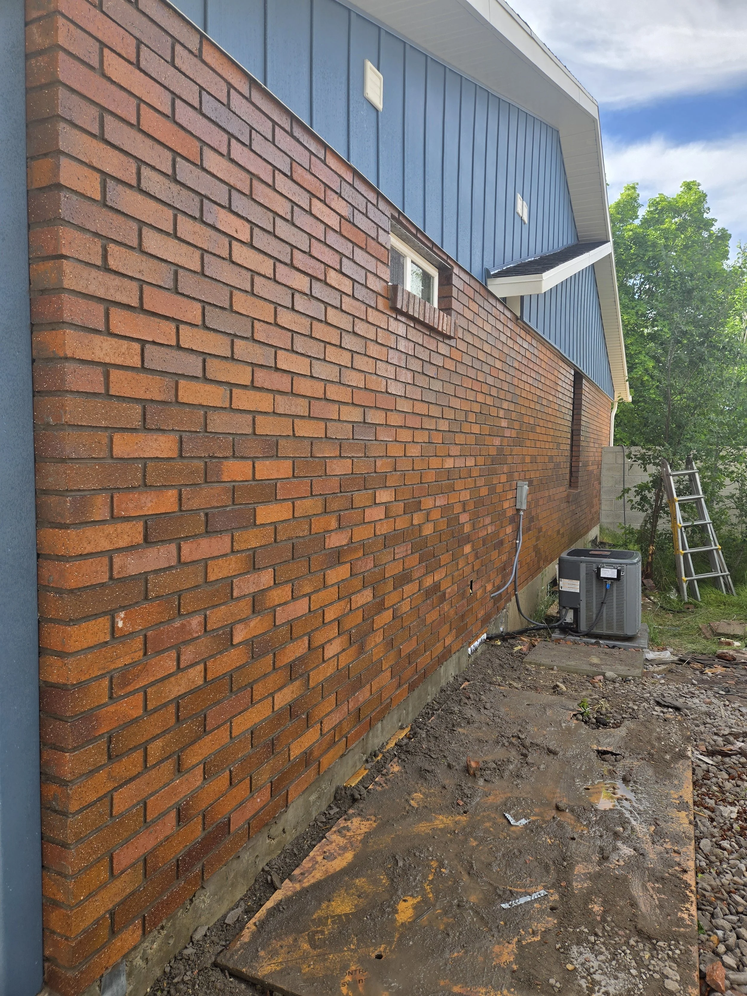 Side of a house with a brick lower exterior and blue upper siding, with a small window, an air conditioning unit, a ladder, and some utility wiring and piping.