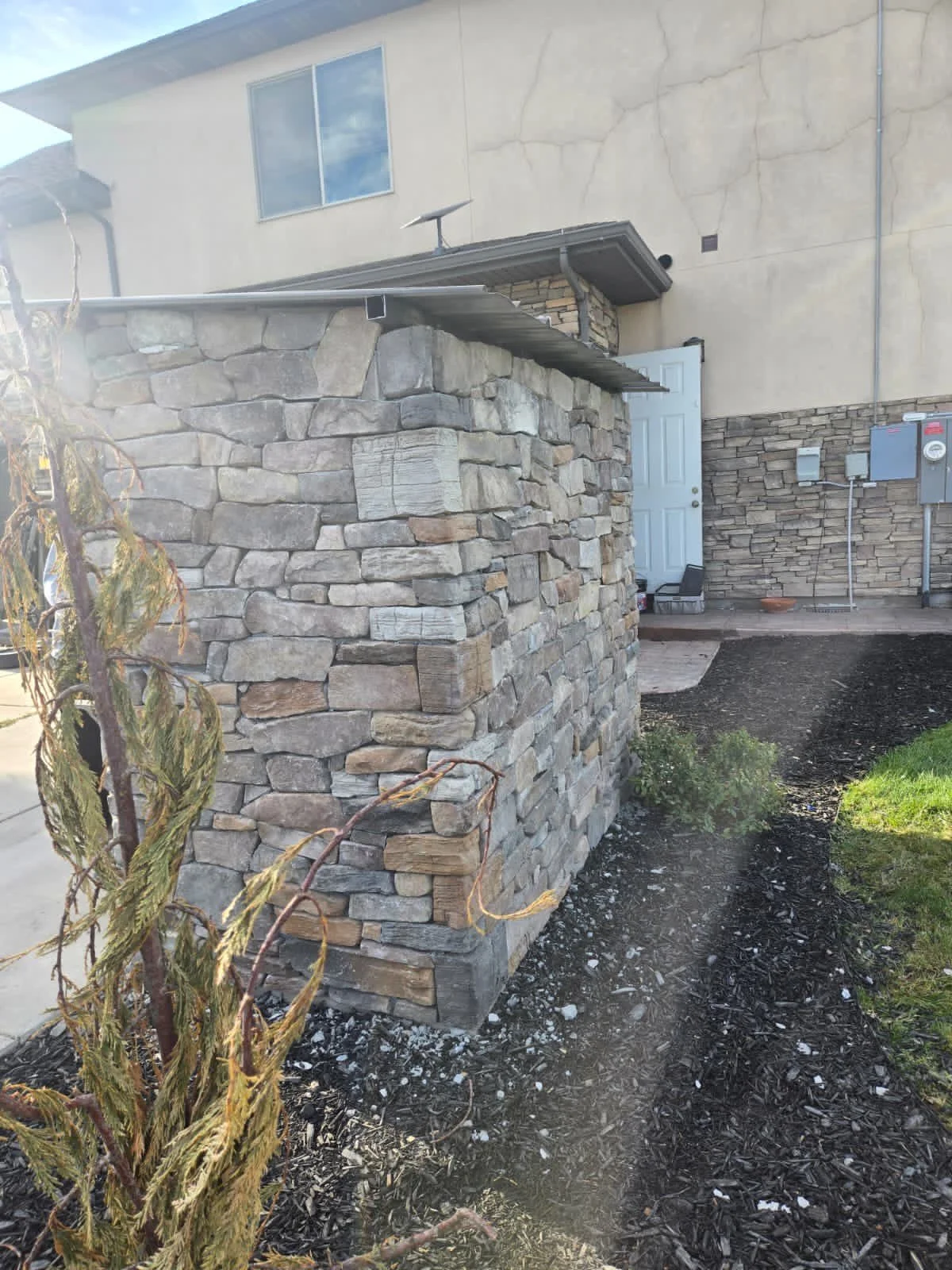 Exterior of a house with a stone wall and landscaping, including a young, dried tree and garden bed with mulch near the house entrance.