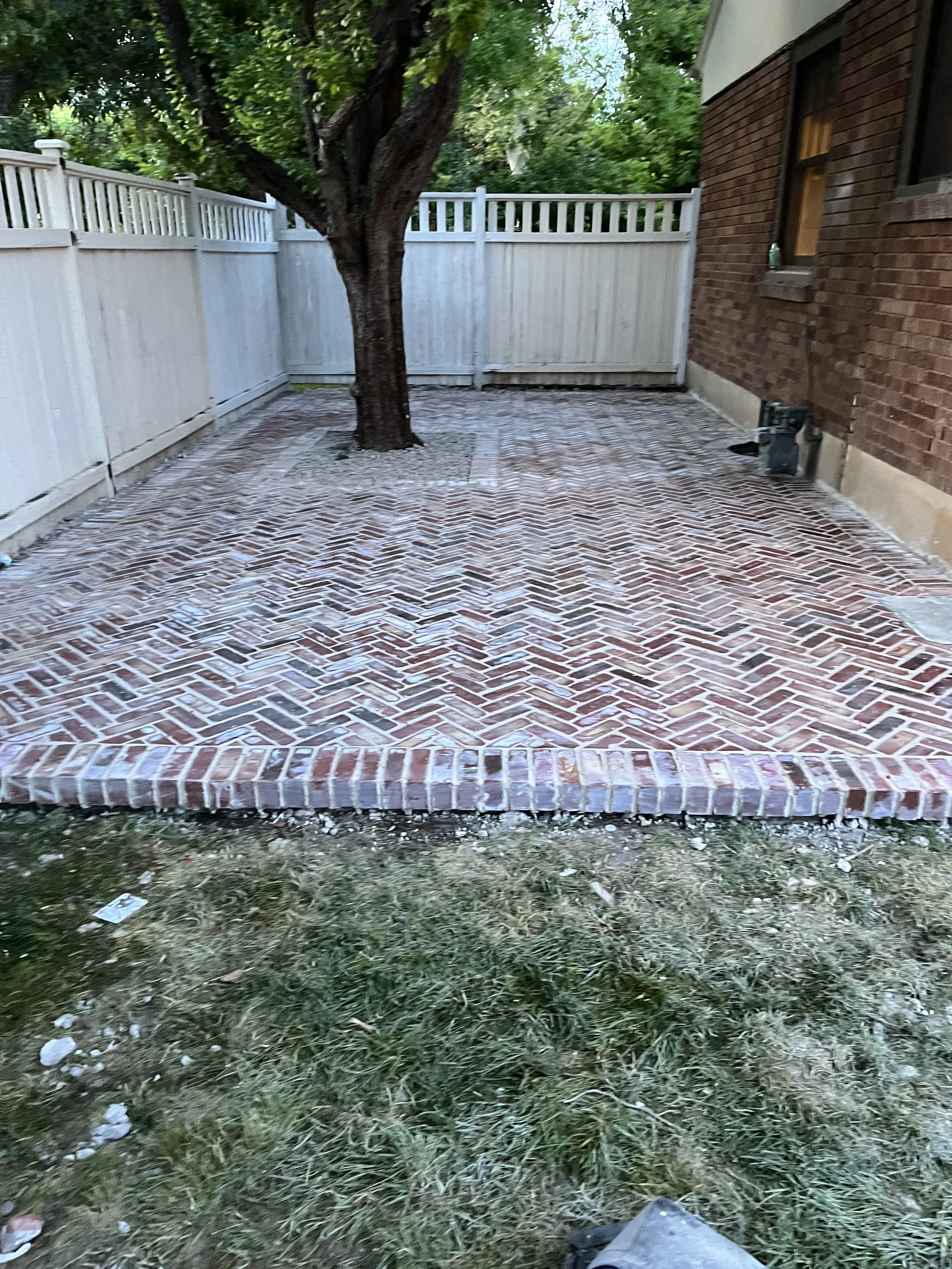 A brick patio with a tree in the center, surrounded by a white wood fence and a brick house on the right side.