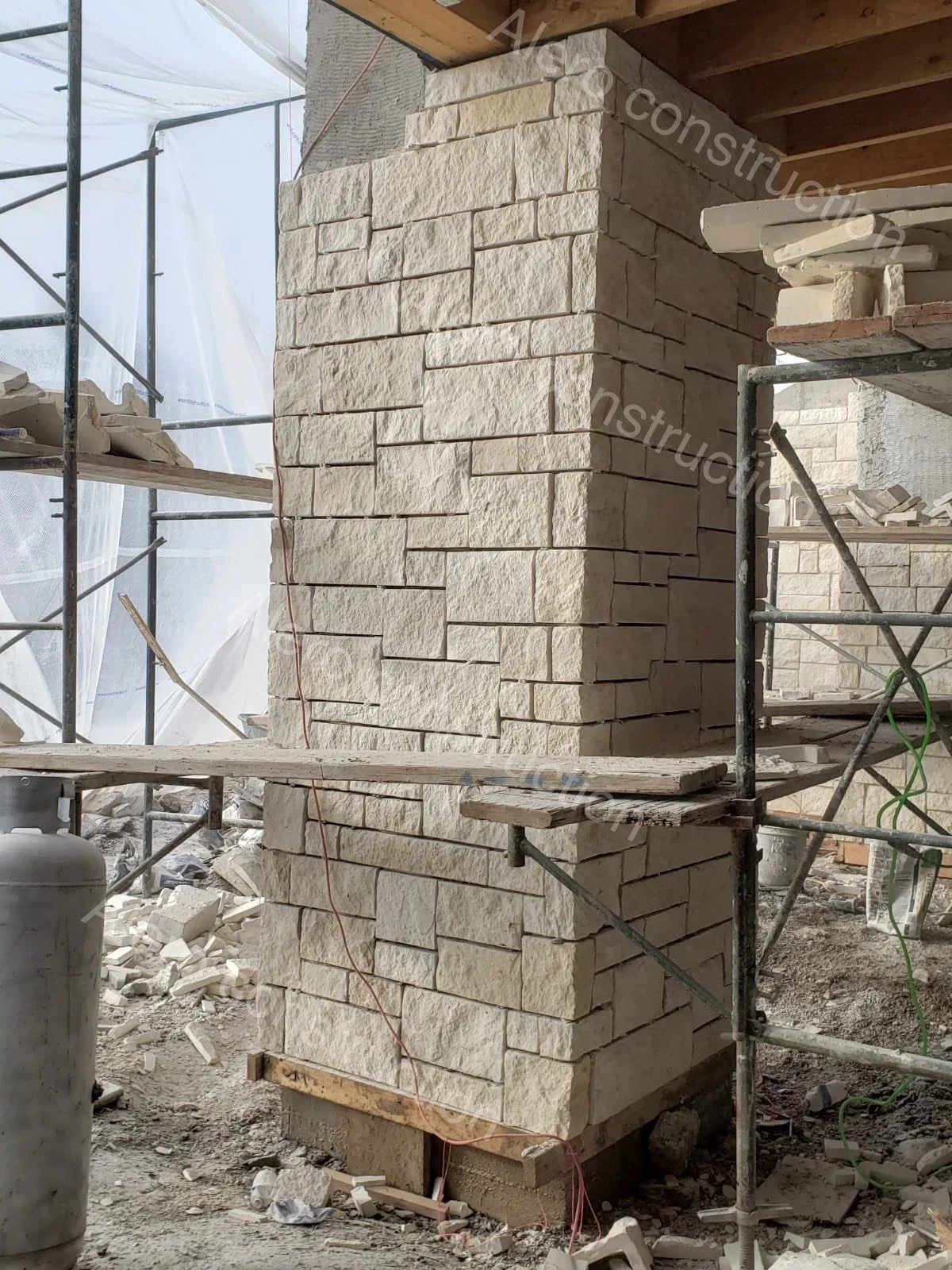 Construction site with a stone brick wall under construction, scaffolding surrounding it, and construction materials on the ground.