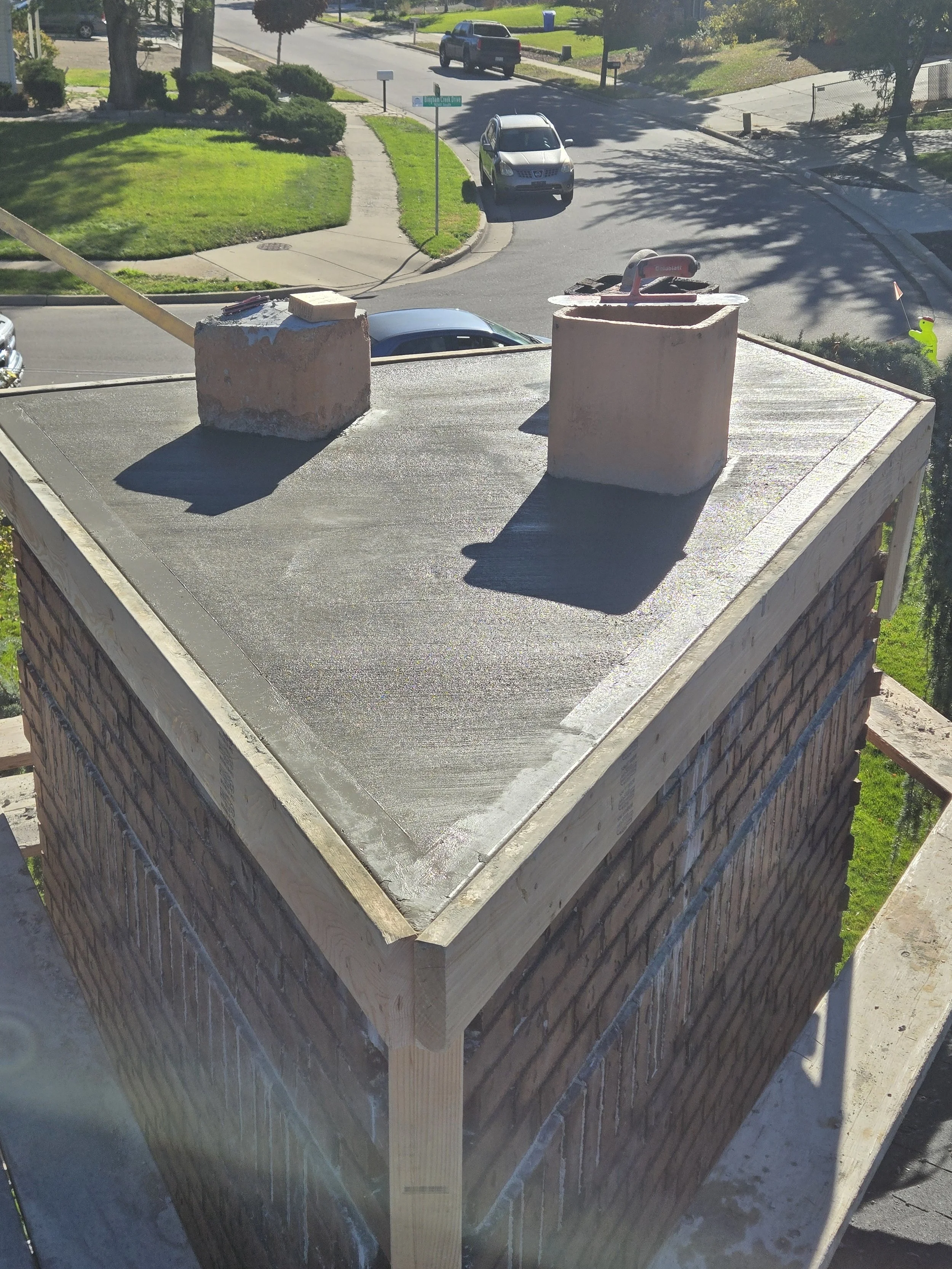 View of the top of a brick chimney under construction with two chimney caps, a trowel, and a brick on the surface, with a residential street and driveway in the background.