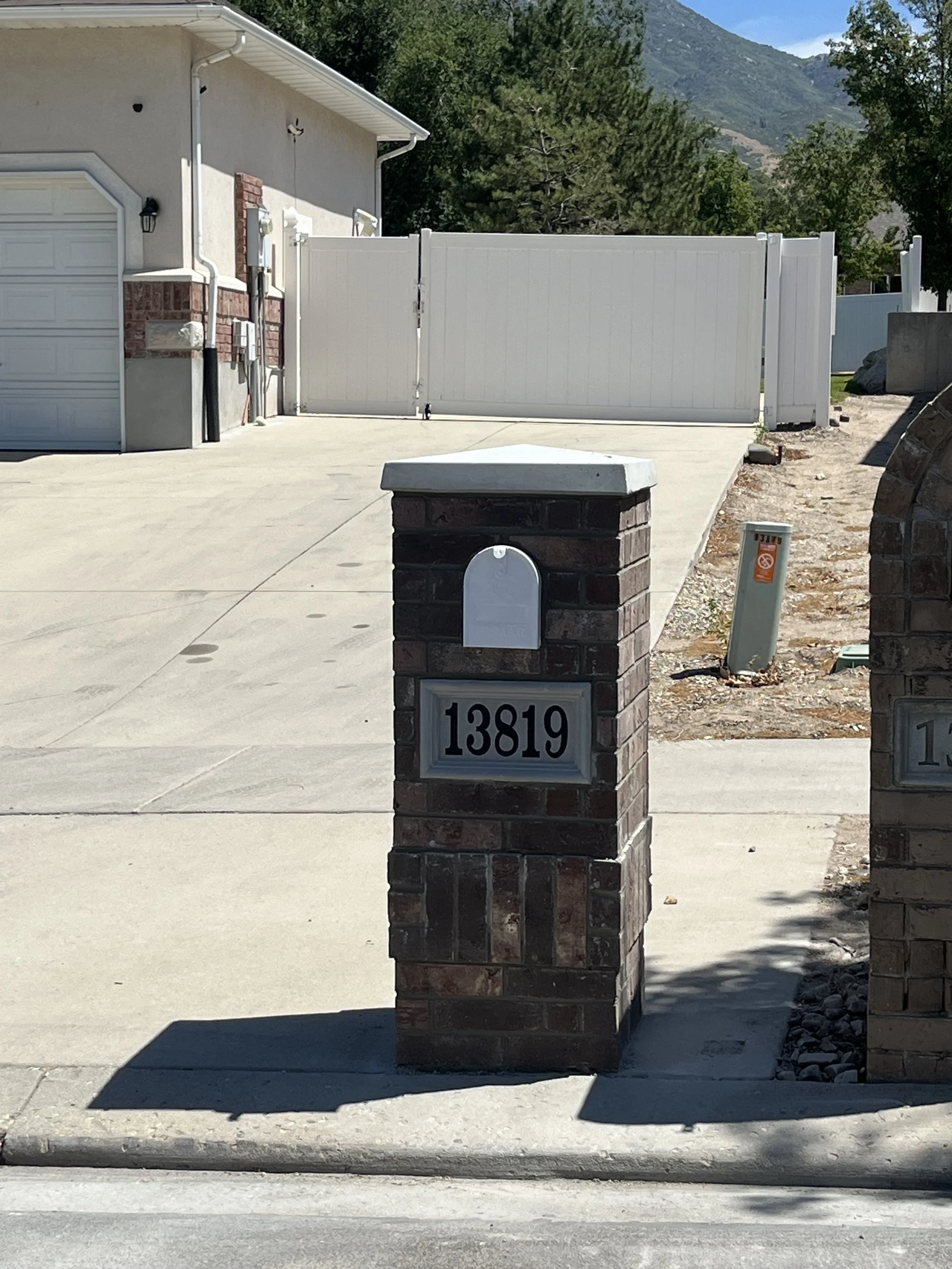 House address number 13819 on a brick mailbox post in front of a concrete sidewalk and driveway, with a white fence and gate in the background, trees, and mountains.