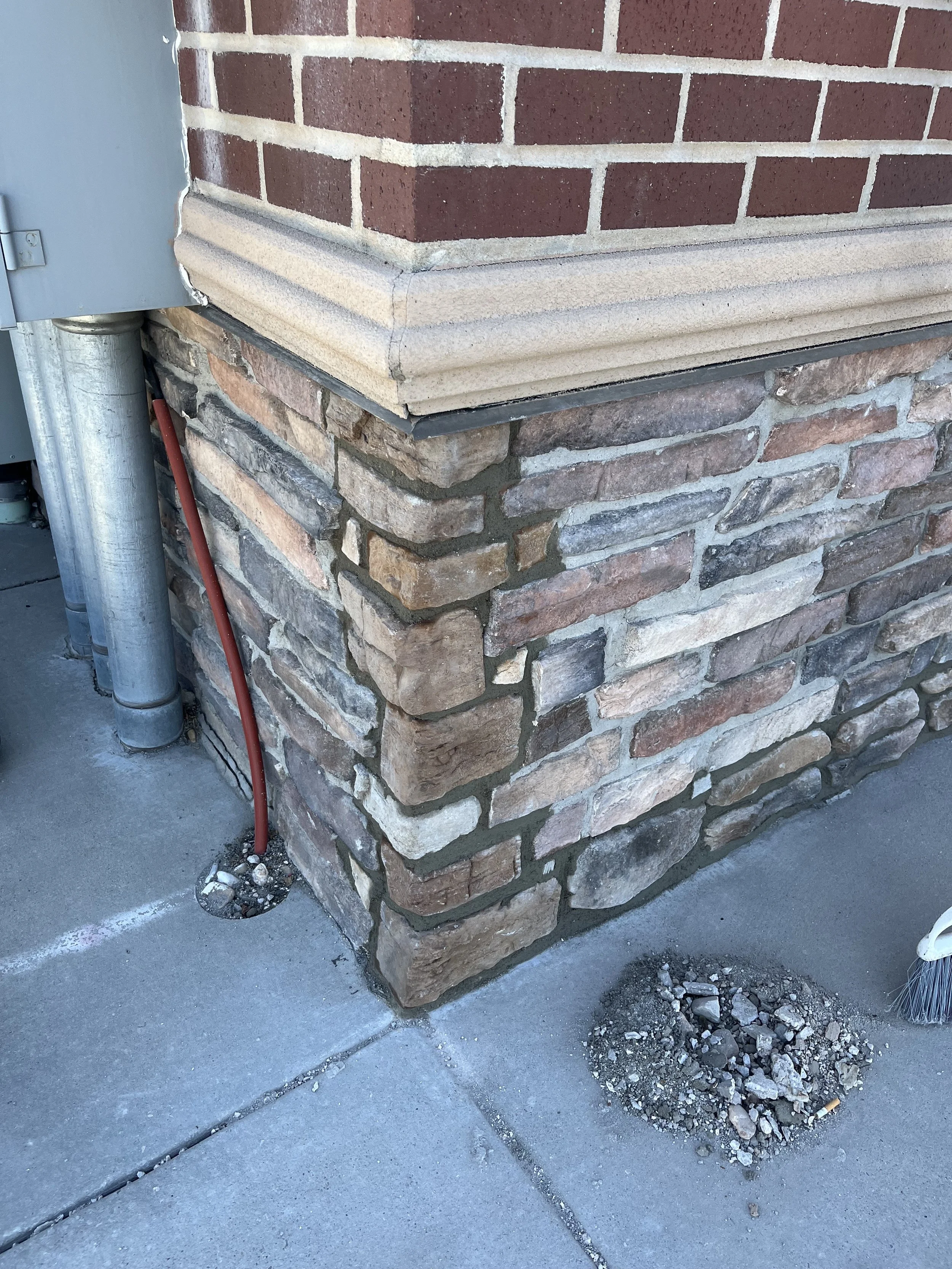 Corner of a brick building with decorative stone base, gravel and debris in a small hole near the corner, a broom leaning against the wall, and an electrical box with a red cable running down to the ground.