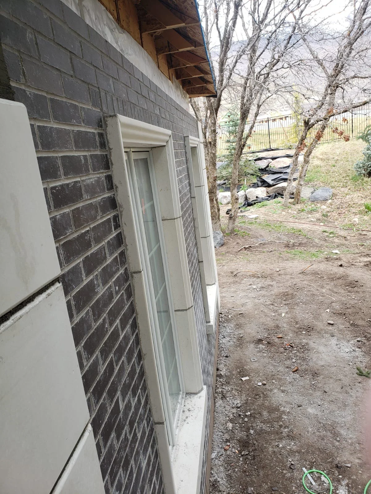 Side view of a house with black brick exterior, white window frames, and unfinished area with exposed wood and roof. In the background, there are trees, rocks, a fence, and a hillside.