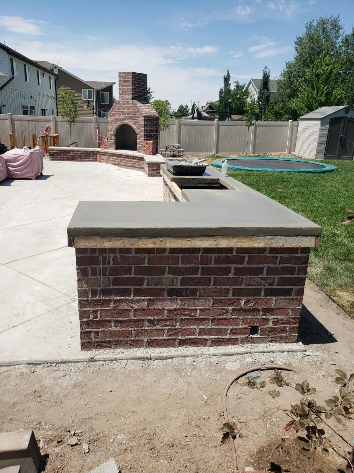 Backyard with a brick fireplace, a concrete counter, and a blue trampoline, fenced yard with trees and neighboring houses.
