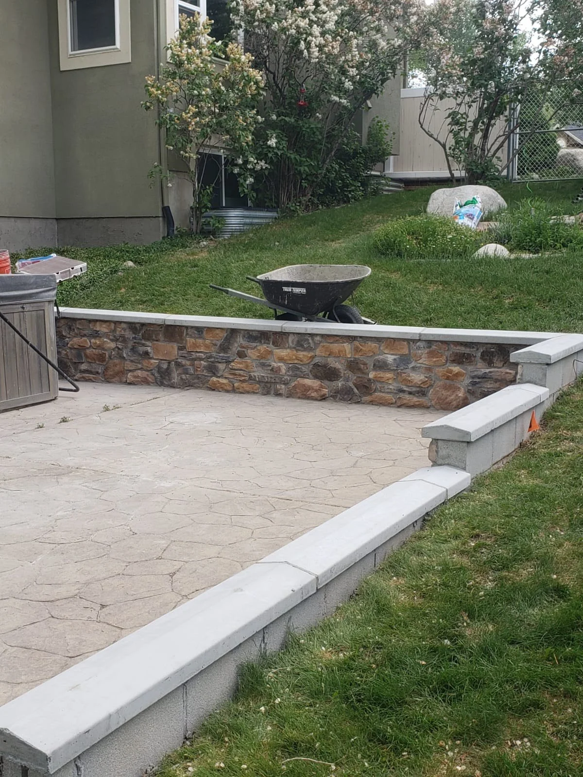 A backyard patio under construction with a stone retaining wall and a wheelbarrow on the grass behind it. There are gardening materials and a shed nearby, with bushes and a flowered tree in the background.