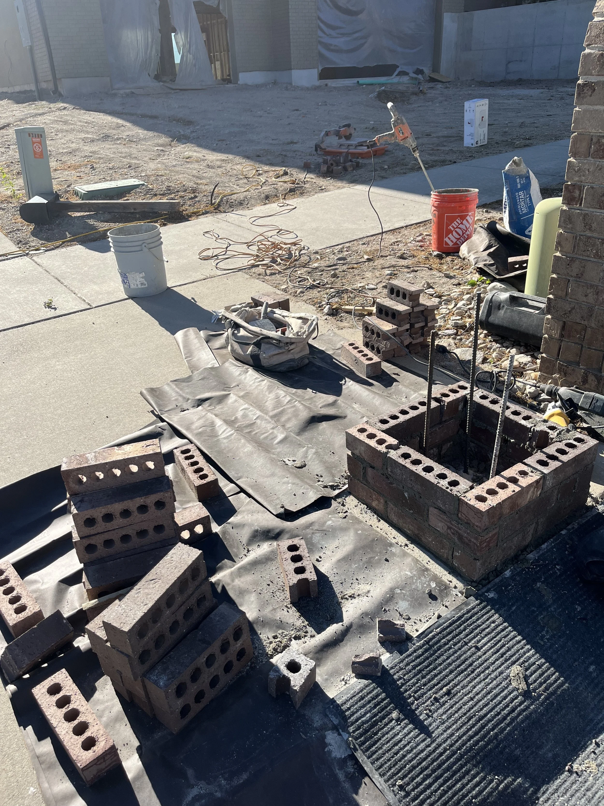 Construction site with bricks, tools, and materials for building a walkway or sidewalk, partially completed with some bricks stacked and others laid down.