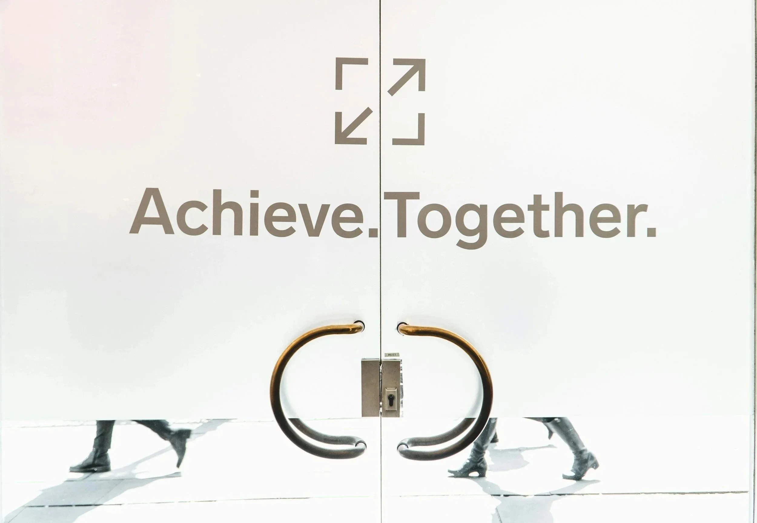 A white double door with the words "Achieve. Together." and a graphic of four arrows pointing outward. The doors have gold door handles, and there are people walking by behind the doors, seen through the glass.