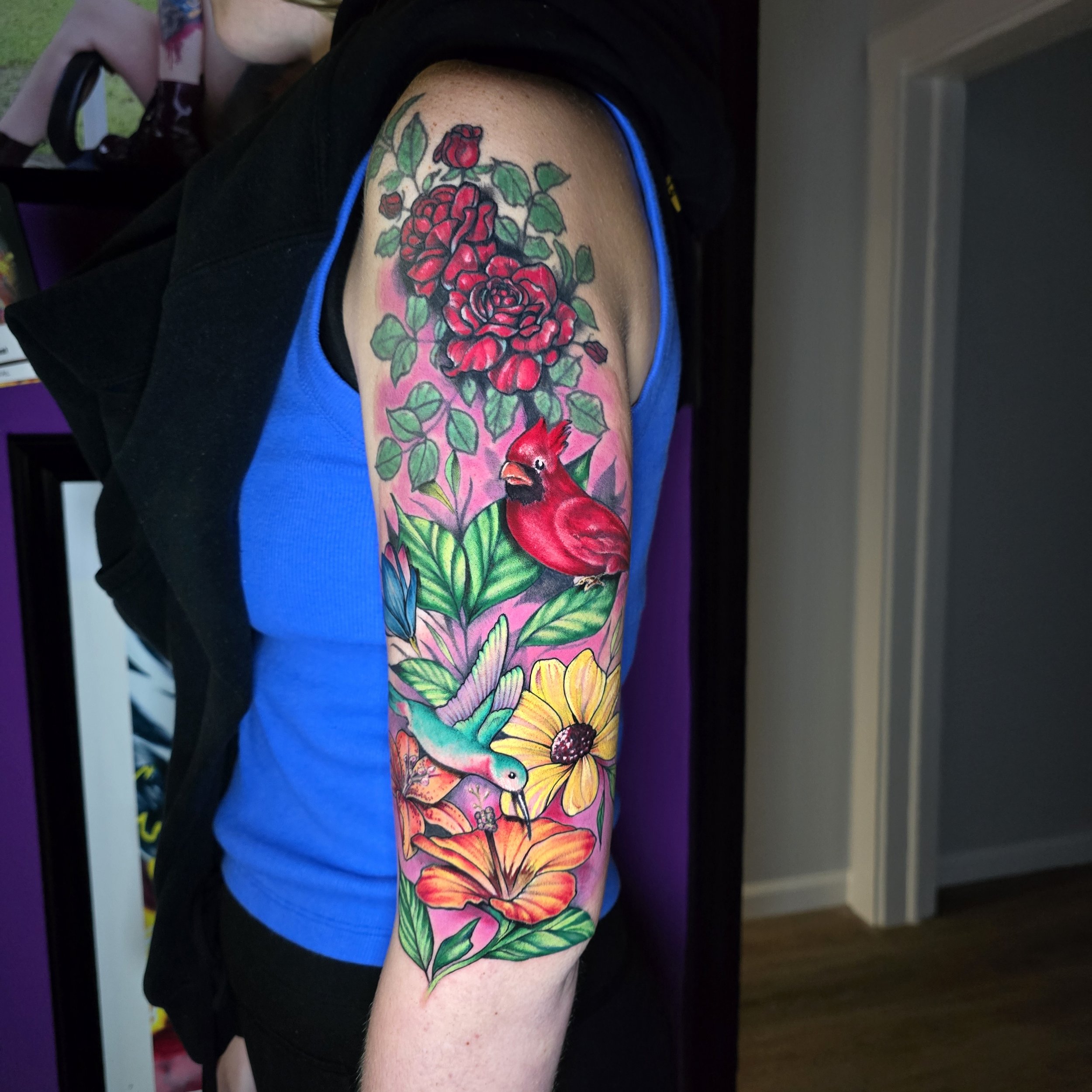 Colorful tattoo of red roses, green leaves, a blue bird, a yellow flower, and a red cardinal on a person's upper arm.
