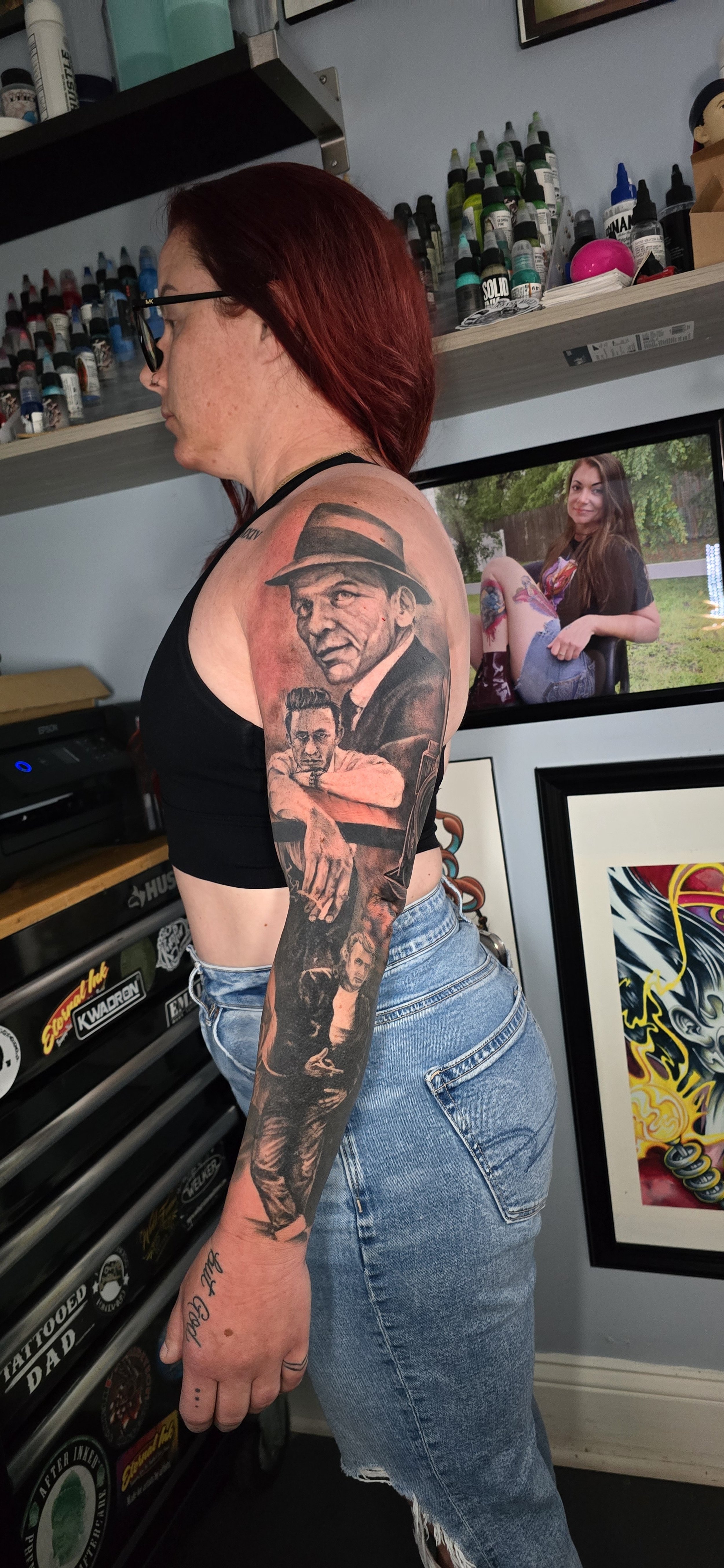 A woman with red hair and glasses shows tattoos on her left arm, featuring portraits of men in black and gray ink. She wears a black crop top and distressed jeans, standing in front of tattoos, artwork, and spray paint bottles.