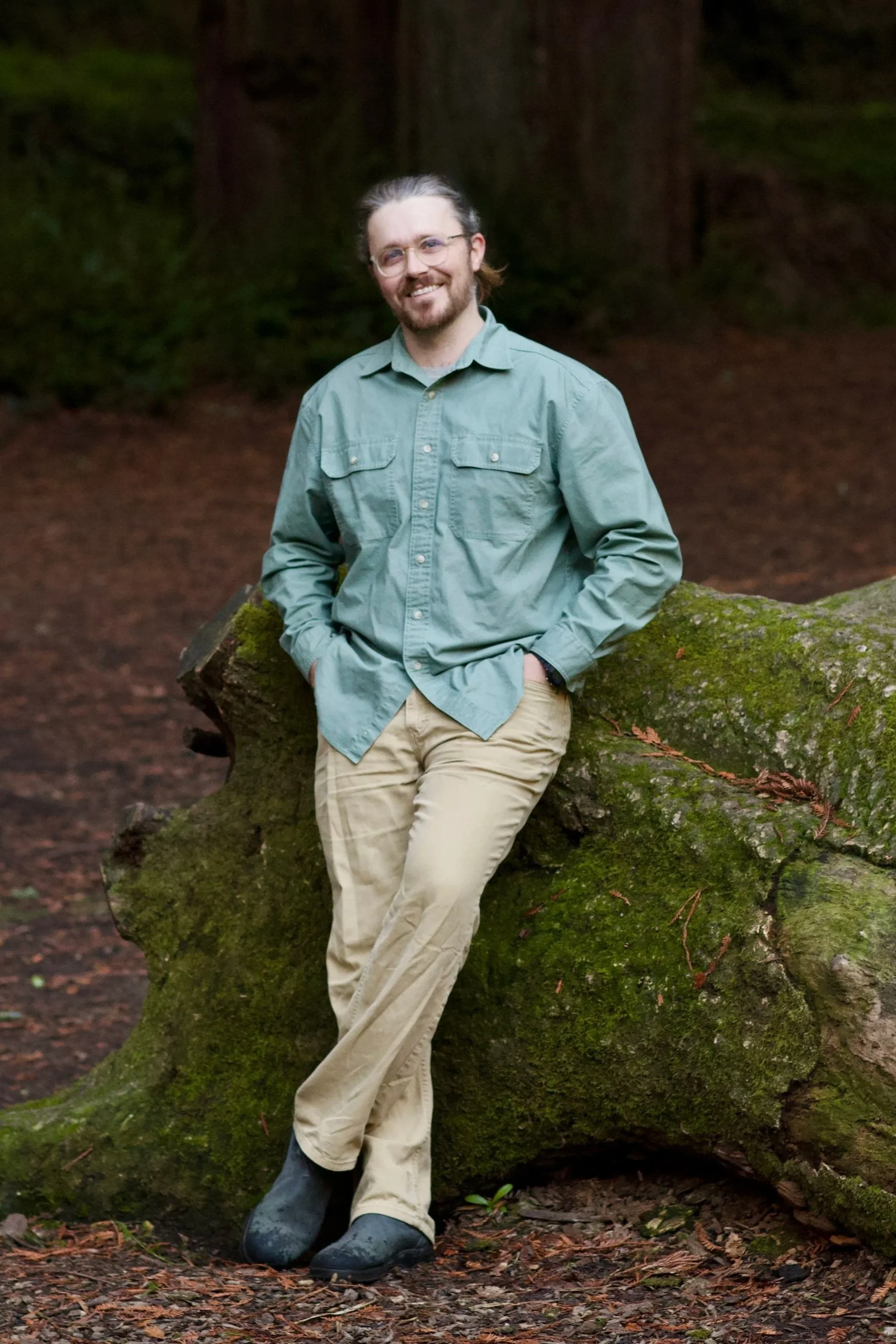 A man with glasses, a beard, and long hair tied back, wearing a green button-up shirt, khaki pants, and black footwear, standing outdoors in a wooded area leaning against a moss-covered fallen tree trunk, smiling at the camera.