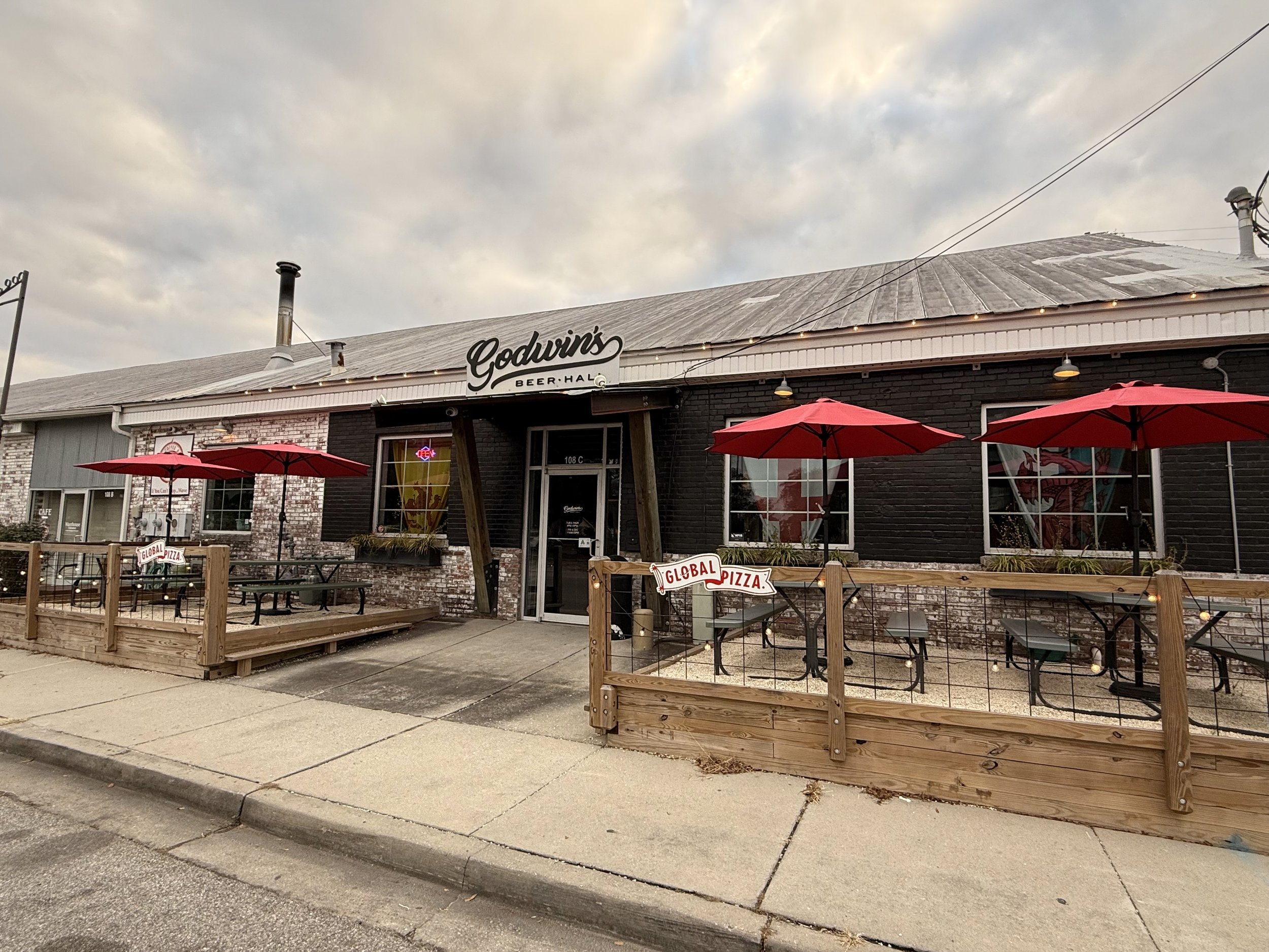 Exterior of Godwin's Beer Hall with outdoor seating area featuring red umbrellas and a sign for Global Pizza.