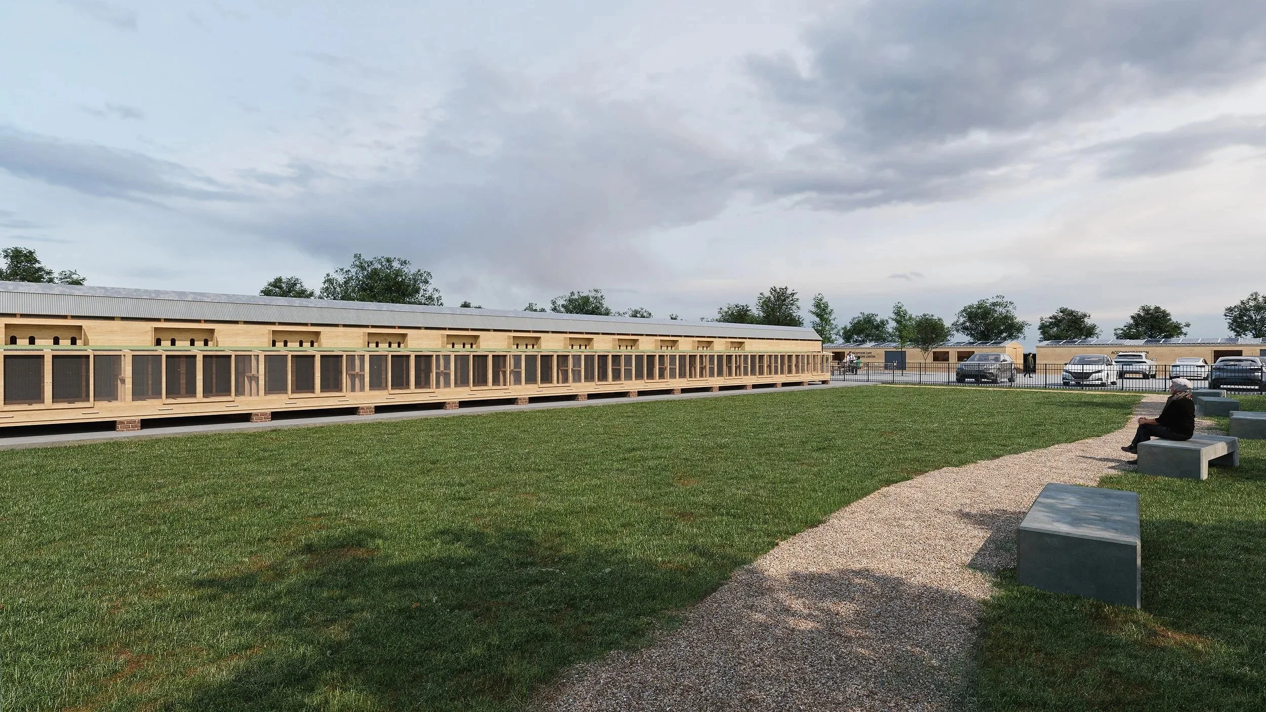 A man sitting on a bench near a grassy area with several rows of dog kennels or cages and parked cars in the background, under a cloudy sky.
