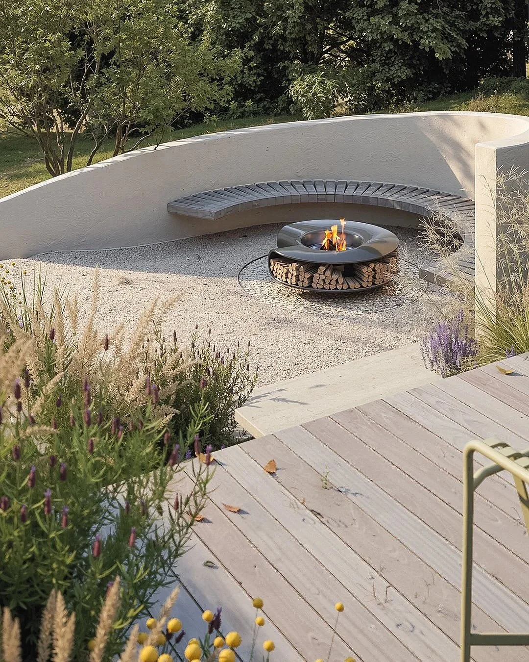 A contemporary outdoor seating vignette featuring a circular fire pit with neatly stacked firewood, a curved concrete bench with timber slats, and light gravel surfacing, framed by soft planting and a warm timber deck to create a premium, modern gard