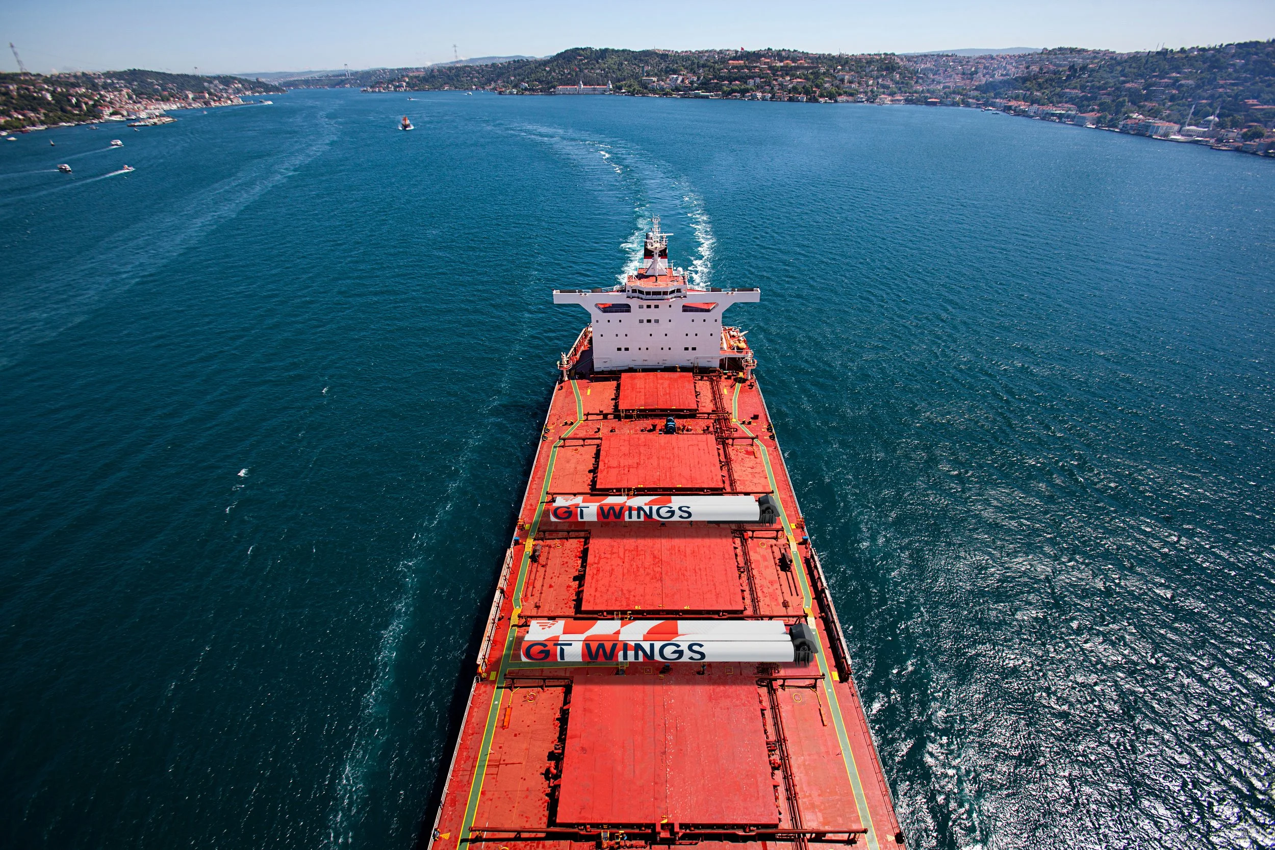 Aerial view of a large cargo ship sailing through a body of water, with a cityscape along the shoreline and green hills in the background.
