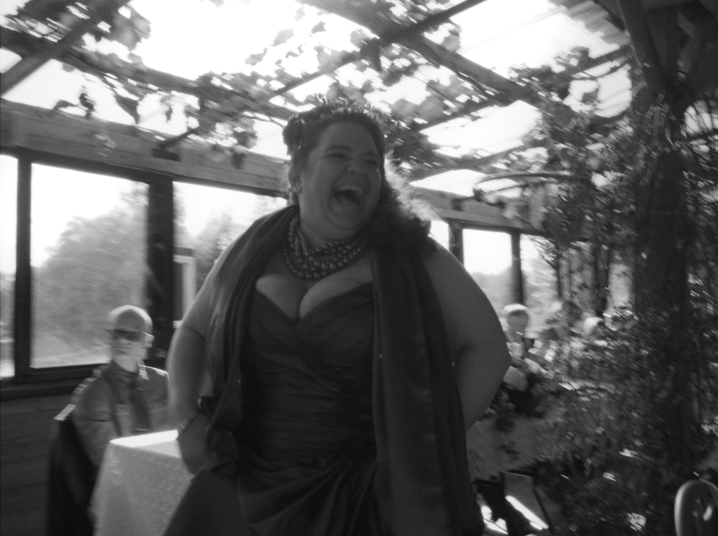A woman laughing and smiling, wearing a necklace and dress, standing outdoors with trees and a person in the background.