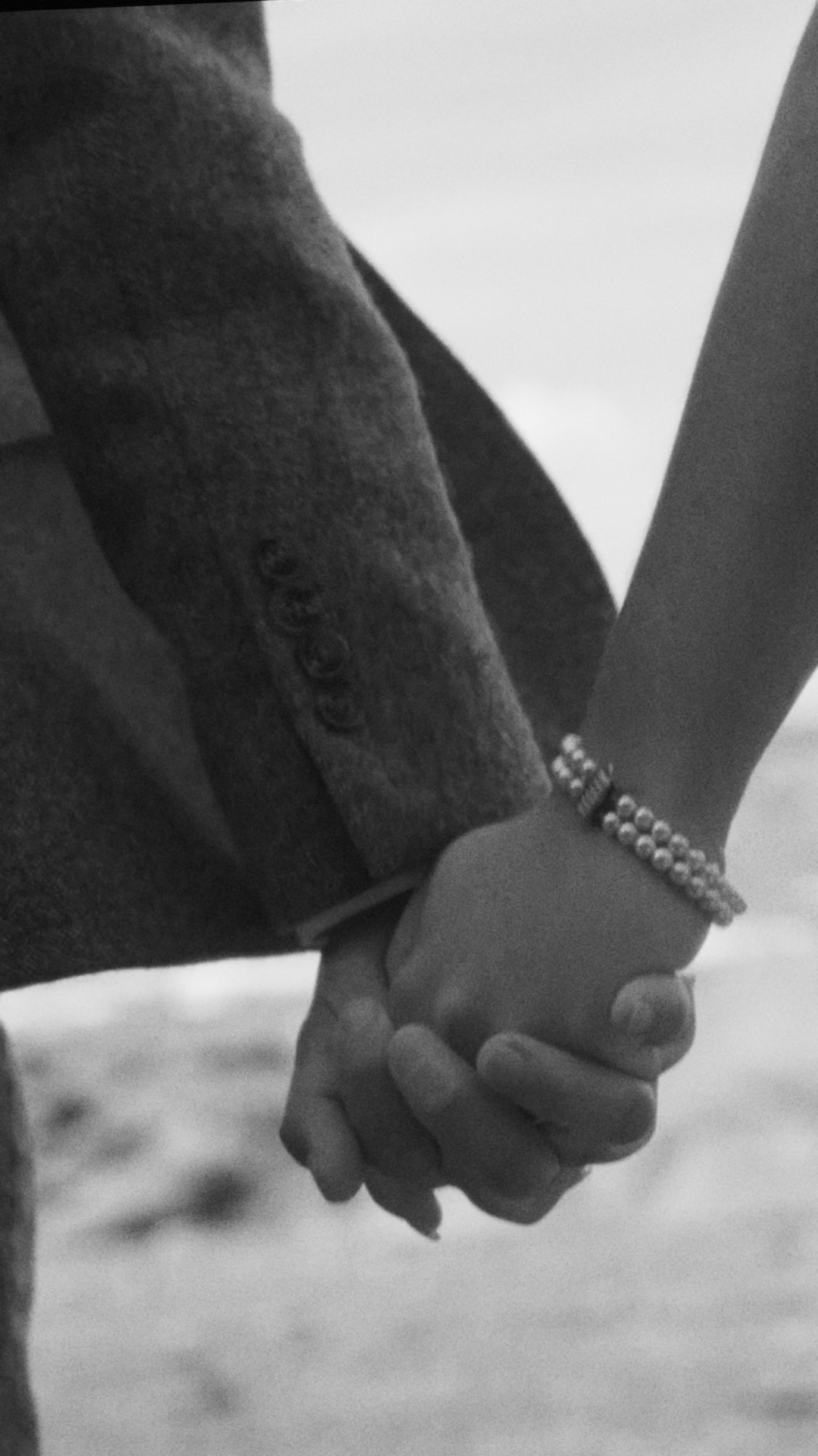 Close-up of two hands holding each other with the person wearing a wristband and a buttoned coat, in grayscale.