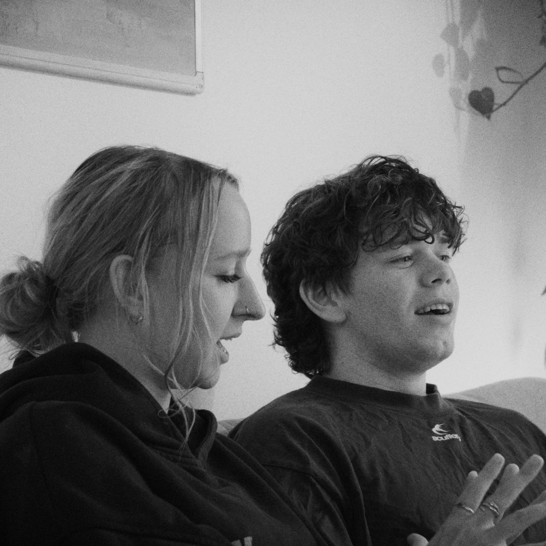 Black-and-white photo of a young woman and a young man sitting on a couch, engaged in conversation, with the woman looking at the man,  with a wall and some decorations in the background.