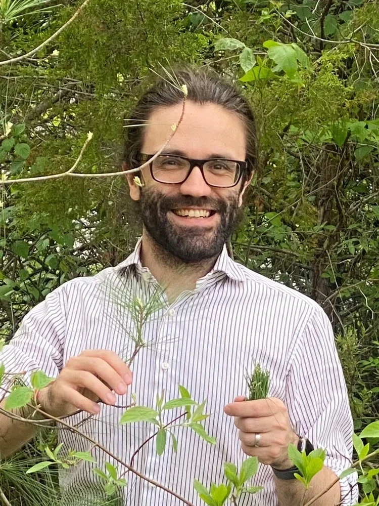 Smiling man with glasses and a beard holding a plant in an outdoor setting surrounded by green bushes and trees.