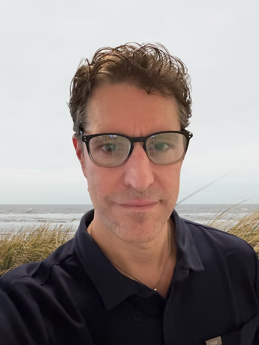 A man wearing glasses and a dark shirt taking a selfie outdoors at the beach on a cloudy day.