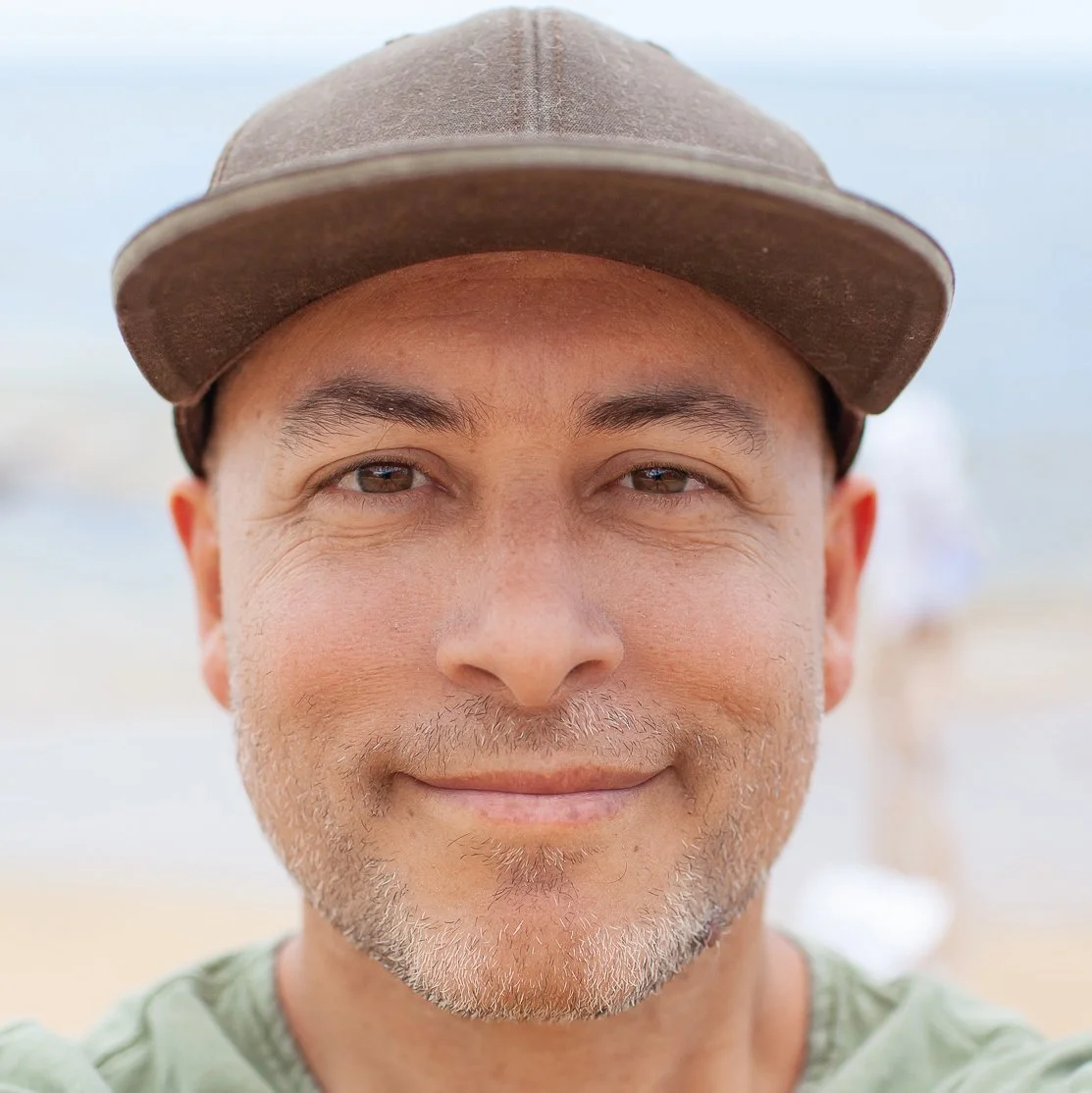 Close-up of a smiling man wearing a brown cap at the beach.