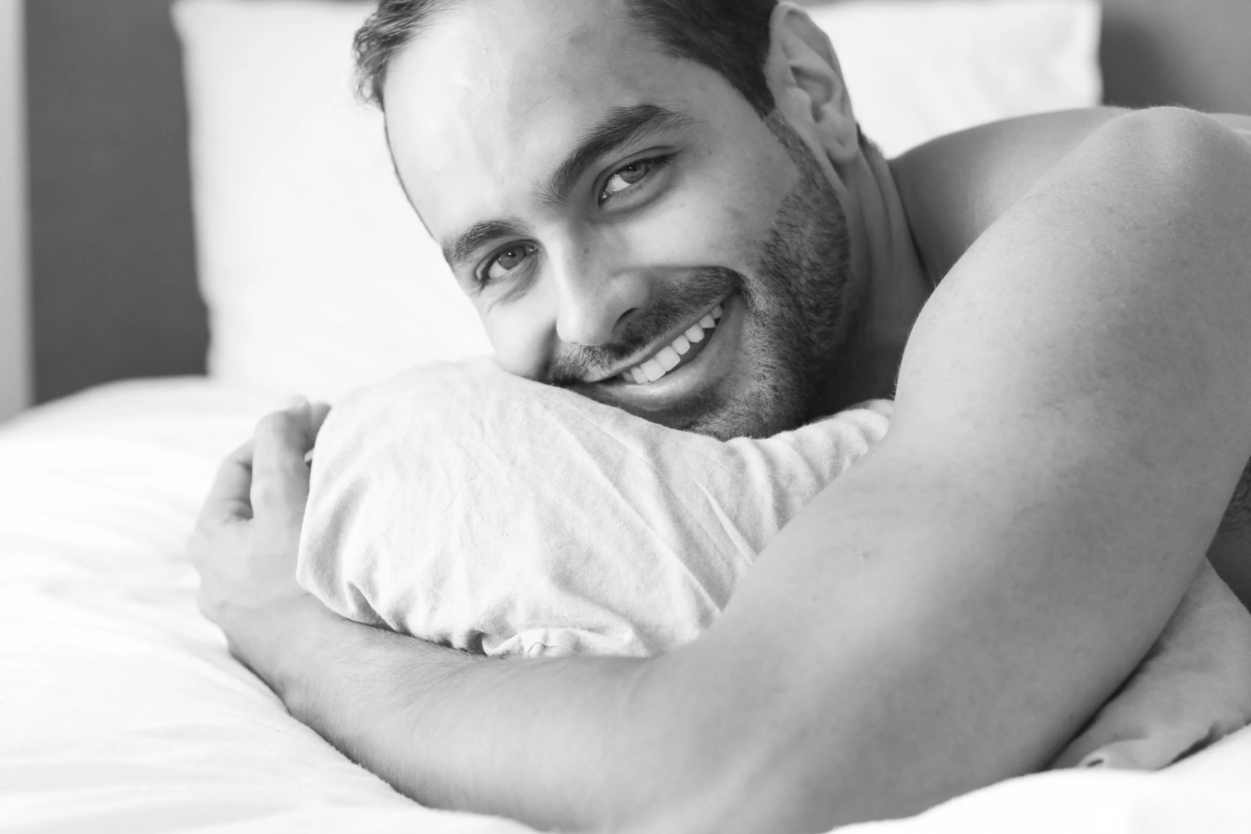 A smiling man with a beard lying in bed, resting his head on a pillow, looking at the camera.