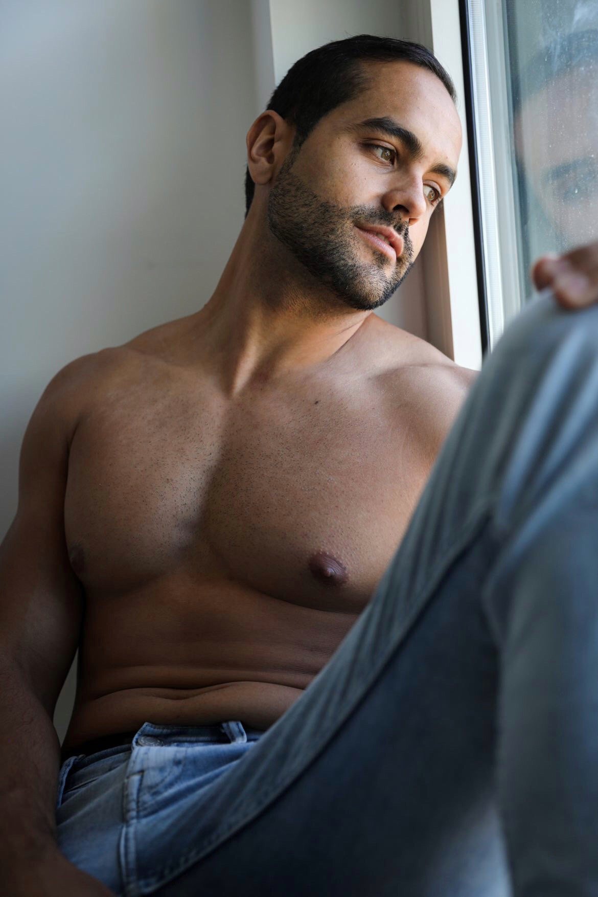 A shirtless man with short dark hair and a beard is sitting by a window, looking out thoughtfully.