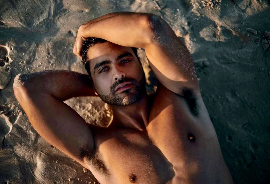 A shirtless man with a beard lying on sandy ground, looking up at the camera with arms resting behind his head.