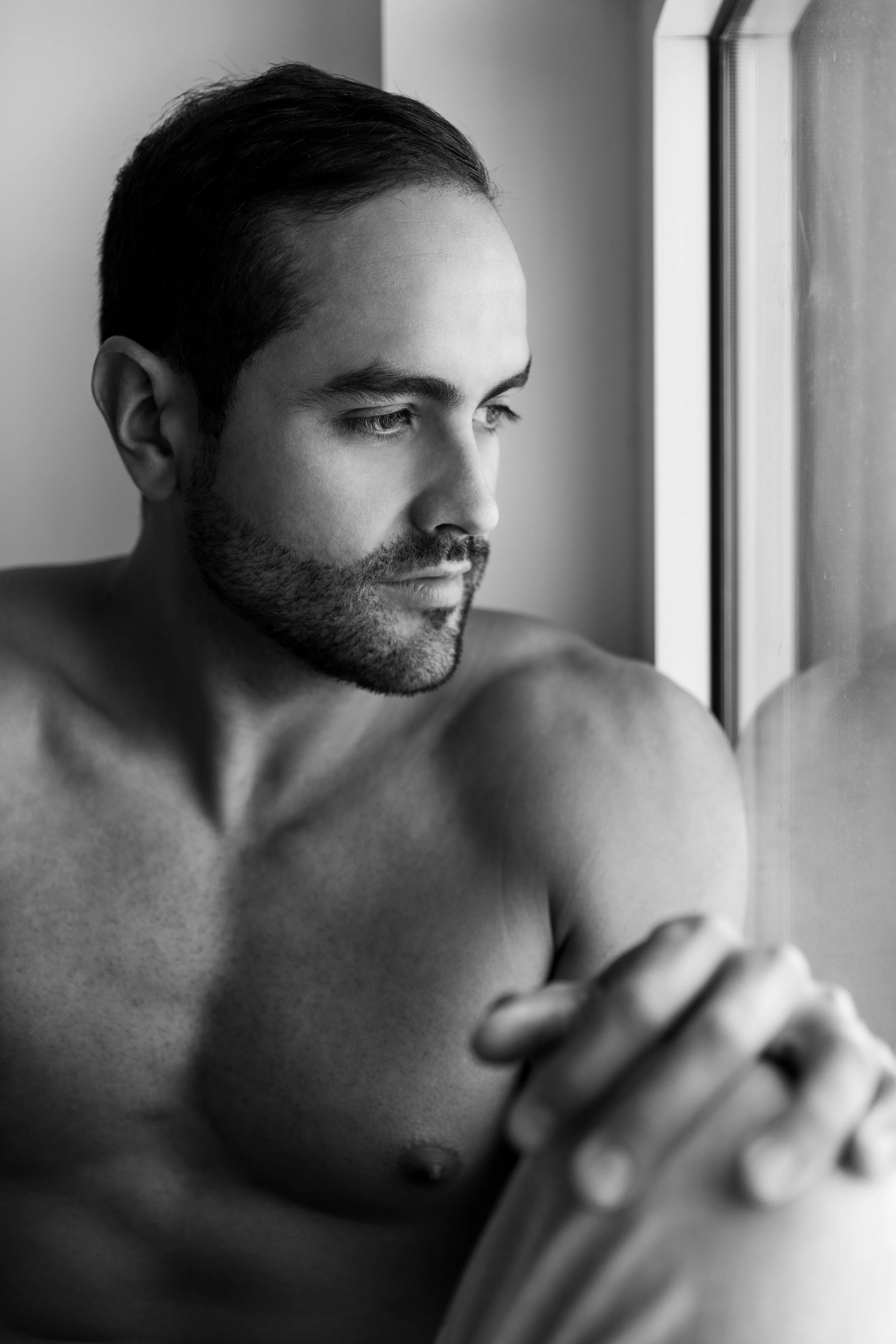 A shirtless man with a beard looking out a window in black and white.