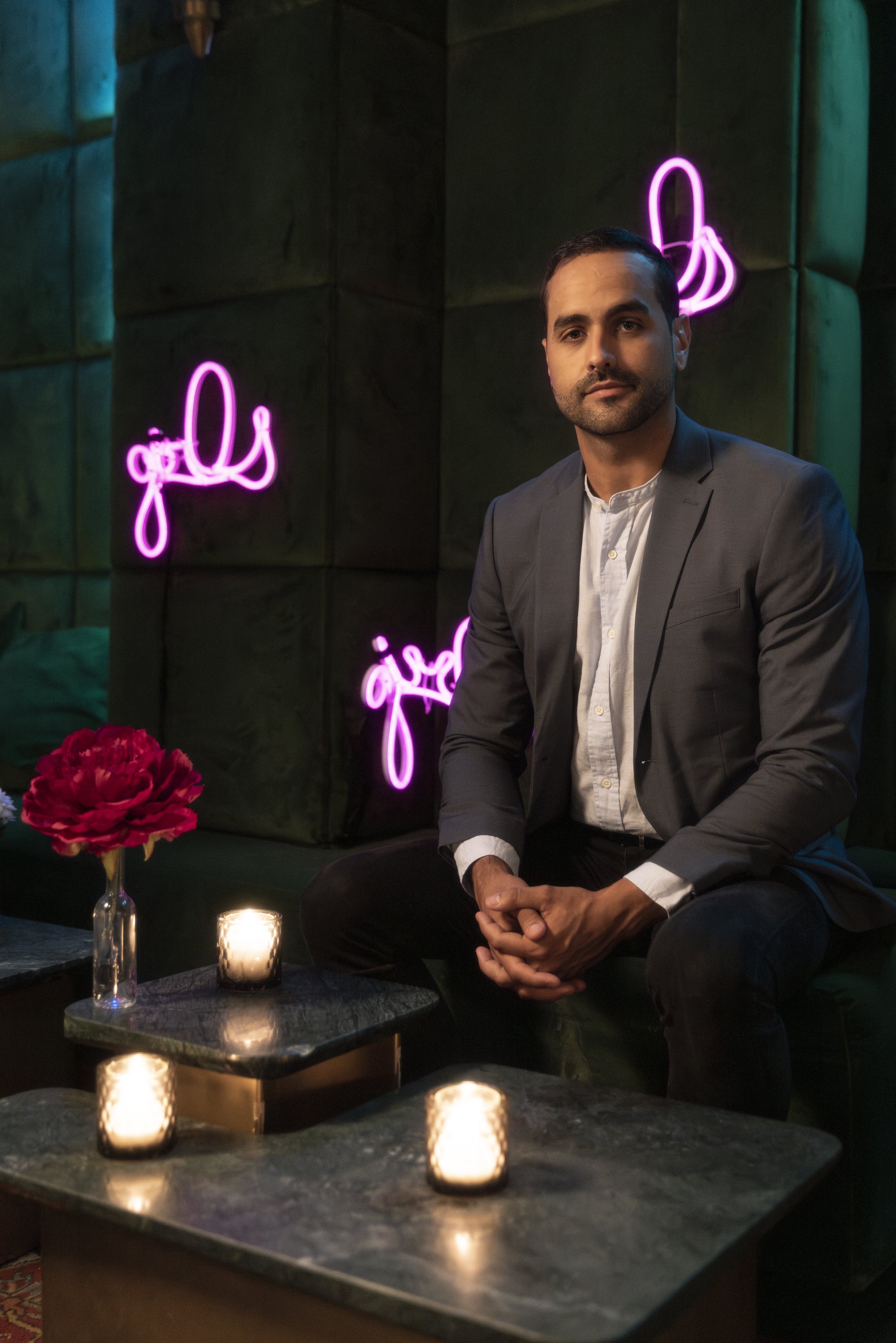 A man in a gray suit sitting on a couch in a dimly lit room with neon pink signs that say 'girls' in the background. There are candles and a vase with pink flowers on a table in front of him.