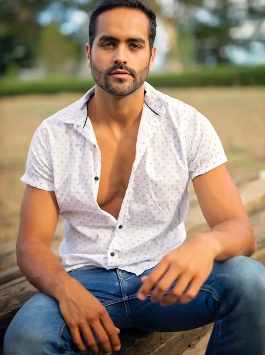 A young man with dark hair and a beard, sitting outdoors on a wooden surface, wearing a white short-sleeve shirt with small pattern and blue jeans, looking directly at the camera.
