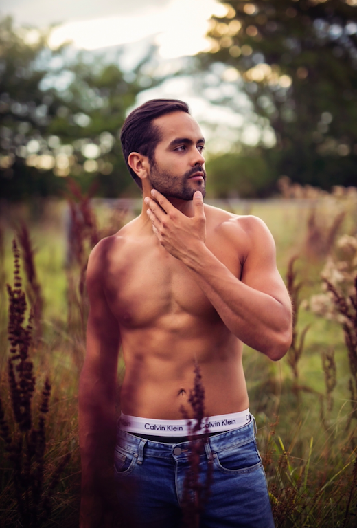 Black and white photograph of a shirtless man with short dark hair and a beard, looking out a window with a contemplative expression.