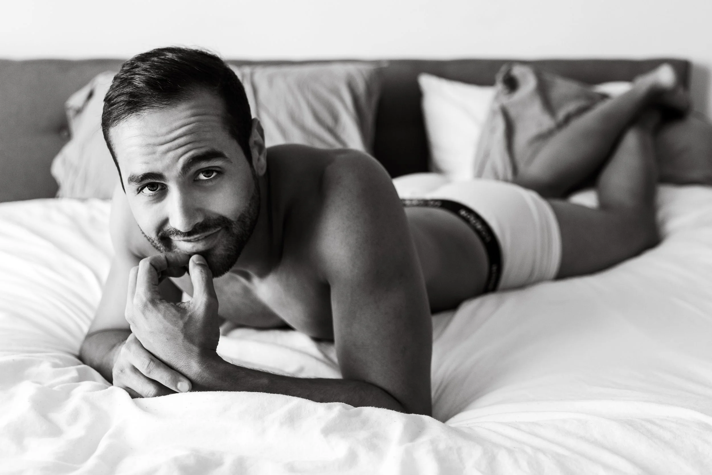 A man with dark hair and a beard lying on a bed, propped up on his elbows, looking at the camera with a slight smile, in a black and white photo.