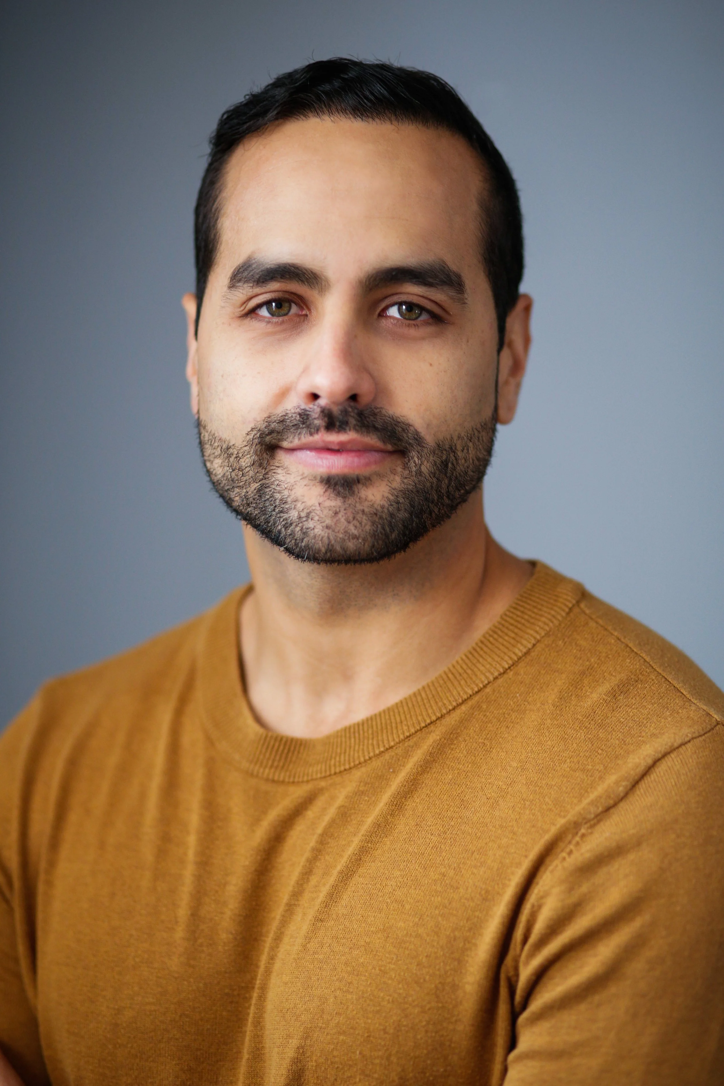 Close-up portrait of a man with dark hair and a beard, wearing a mustard-colored sweater, against a neutral background.