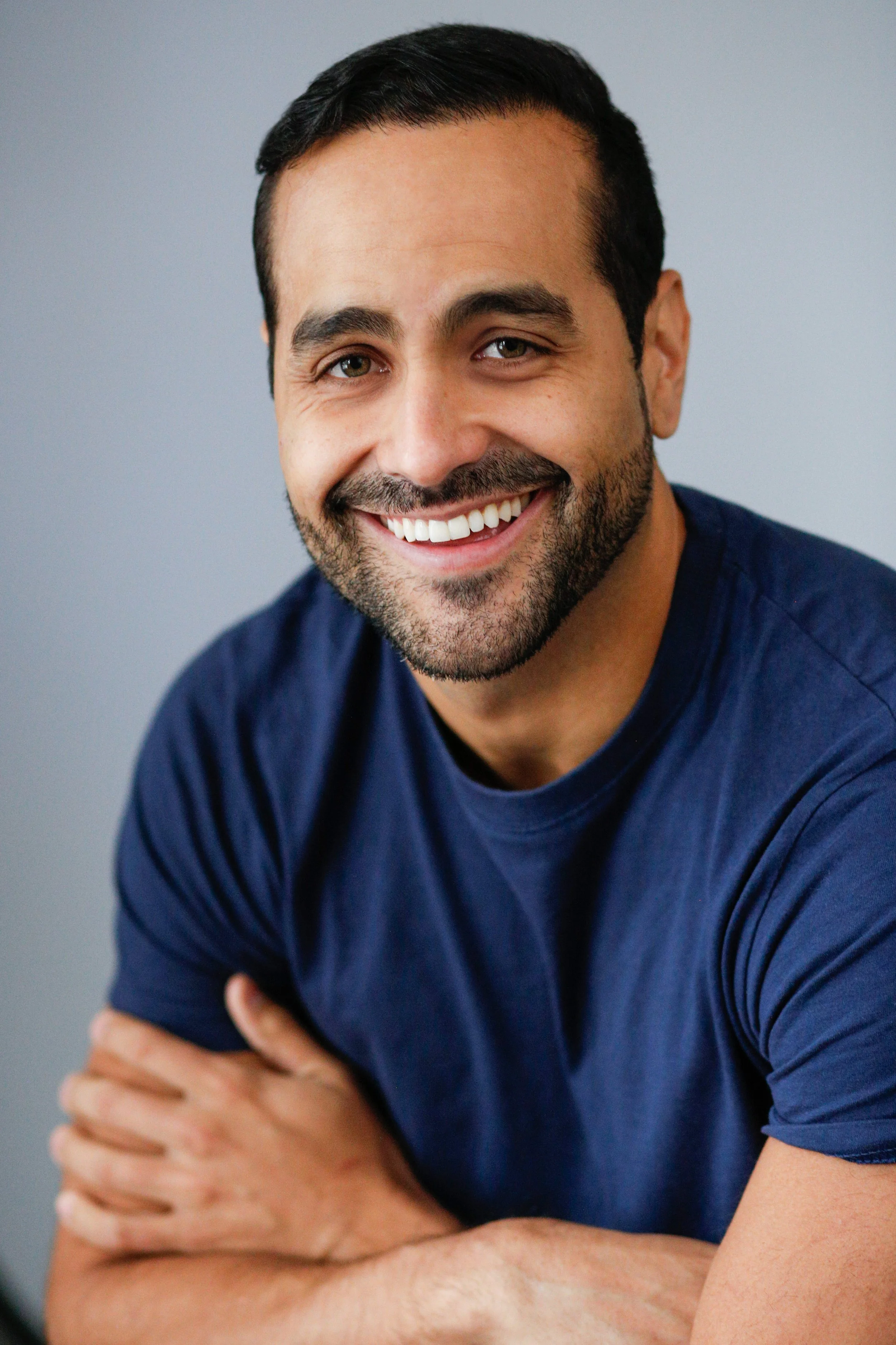 Close-up of a smiling man with dark hair and a beard, wearing a navy blue T-shirt, against a plain gray background.