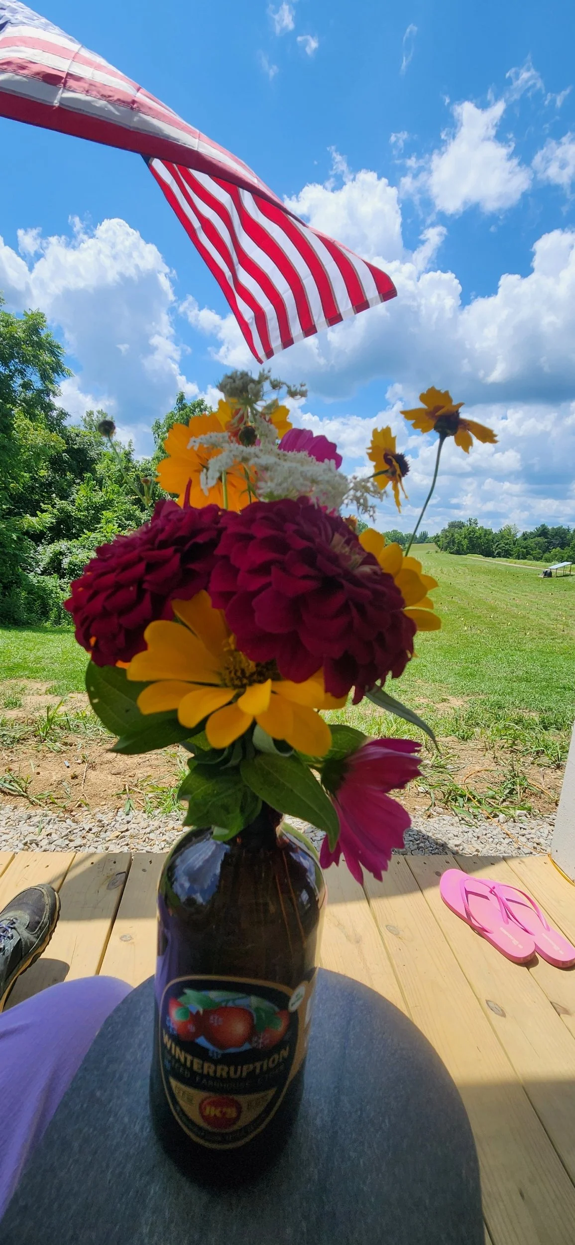 A colorful bouquet of flowers in a glass vase, placed on a wooden deck, with an American flag flying in the background against a partly cloudy blue sky.