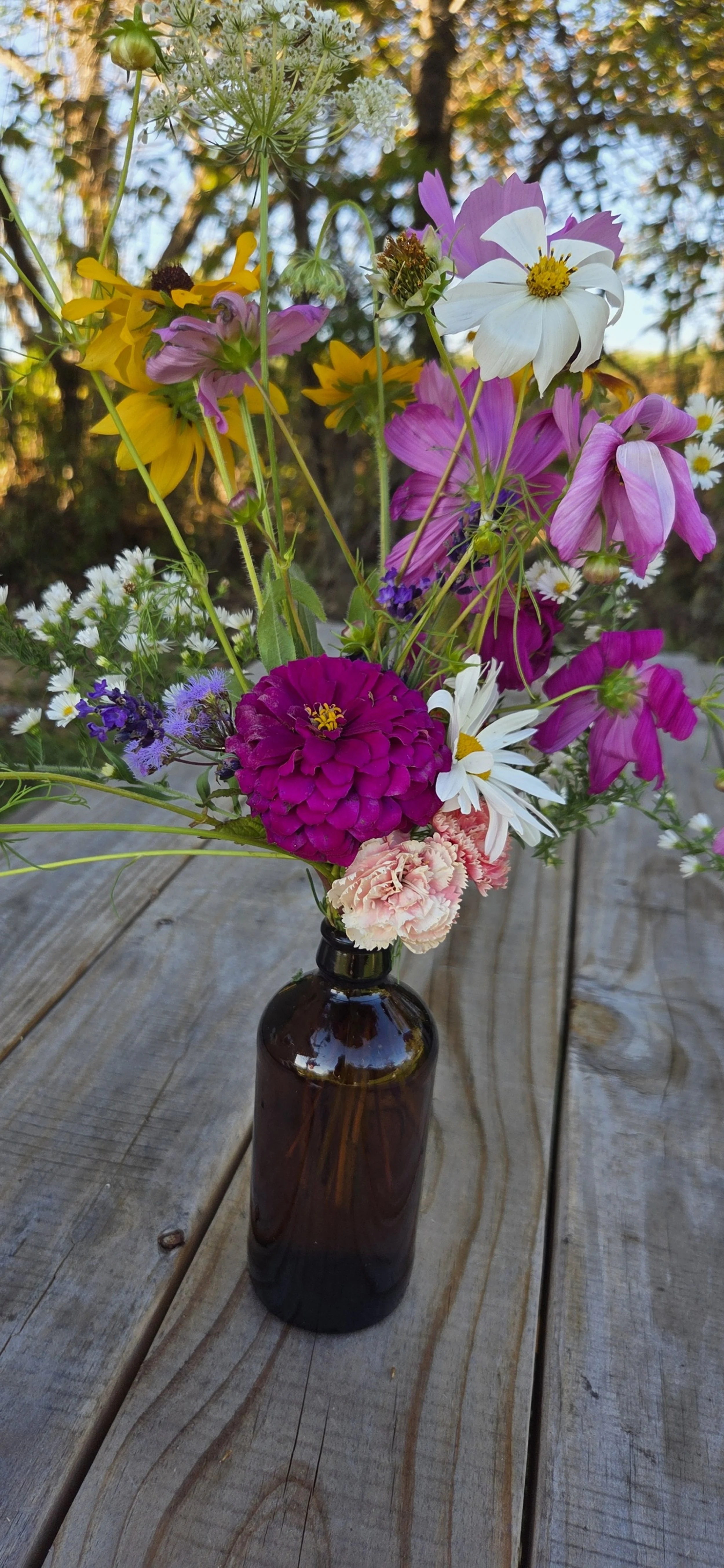 A bouquet of colorful flowers in a small brown glass vase placed on a wooden surface outdoors.