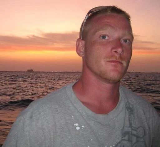 A man with short hair and sunglasses on his head standing near the ocean at sunset, with a cloudy sky and a distant building on the horizon.