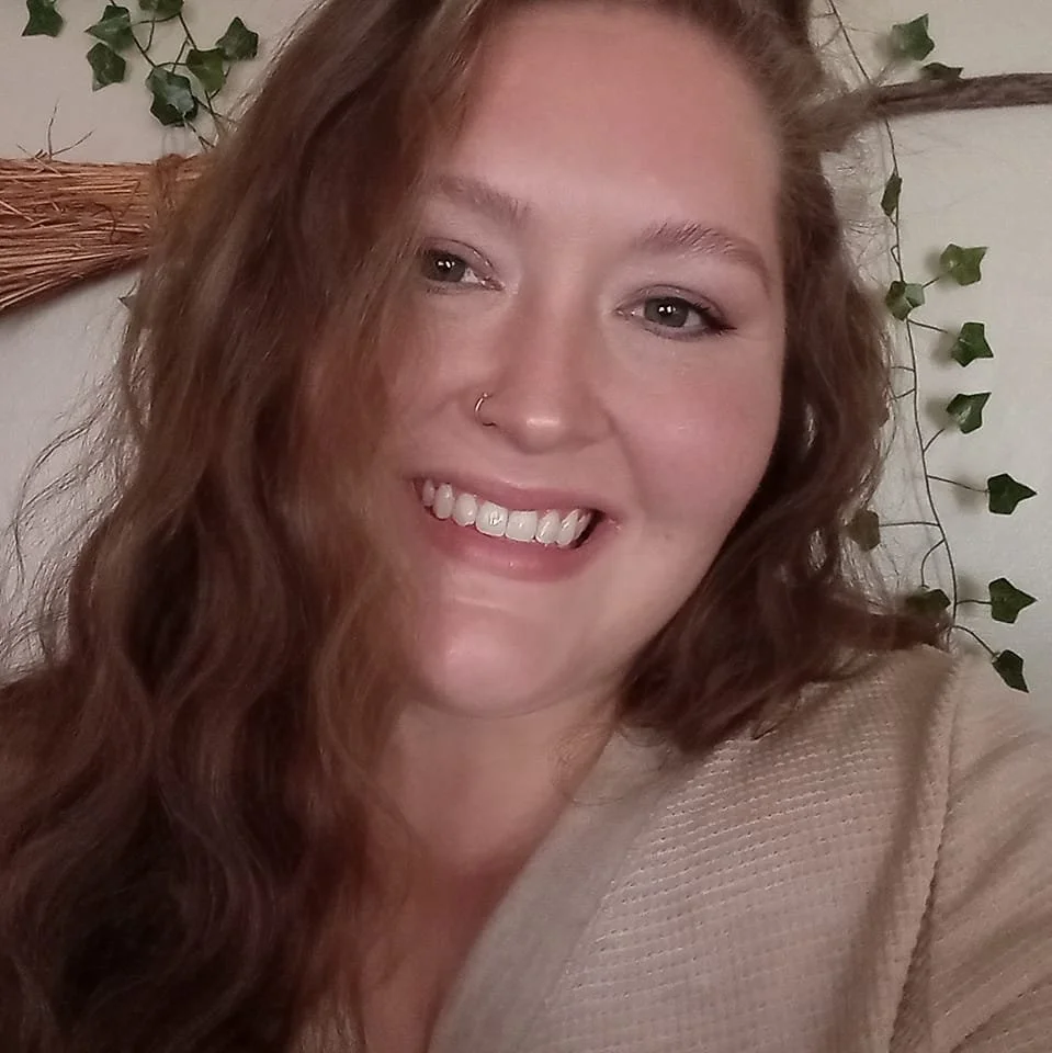 A smiling woman with long, wavy red hair and a nose ring, posing in front of a wall decorated with small greenery and a wicker basket.