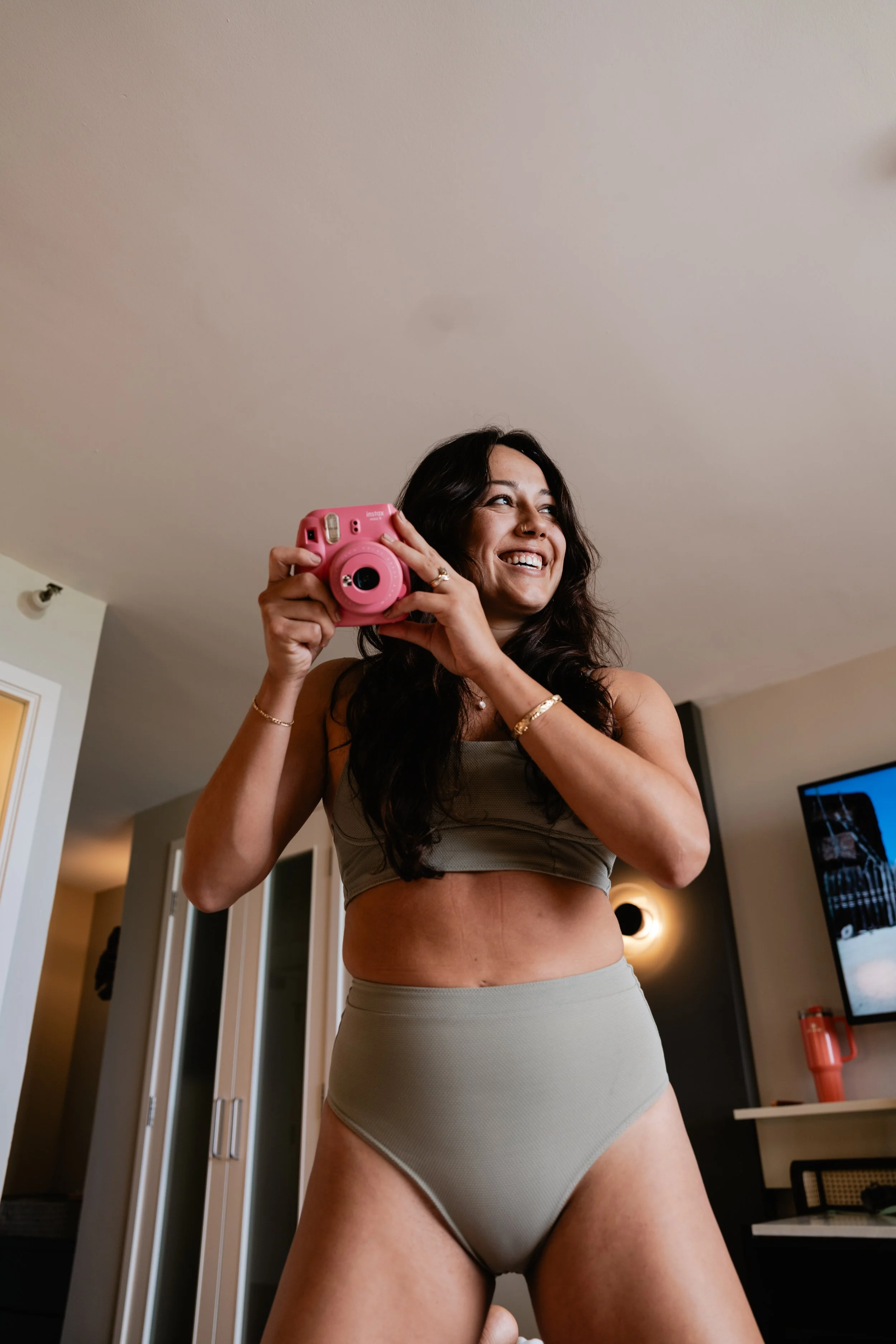 A woman with dark hair, wearing beige underwear, holding a pink instant camera, smiling in a modern living room.