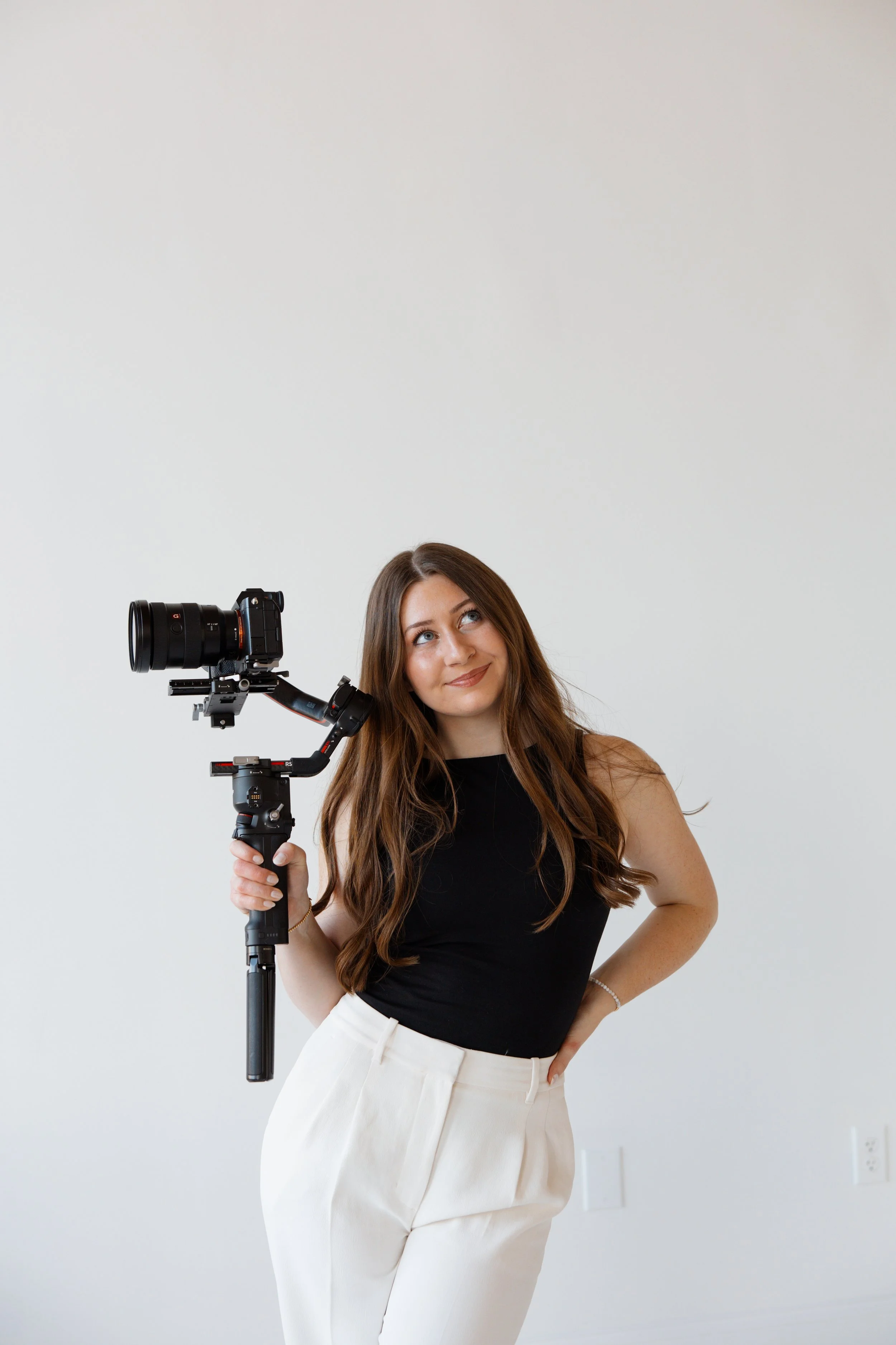 Videographer + Photographer, A woman holding a camera stabilizer with a camera attached, standing against a plain white wall.