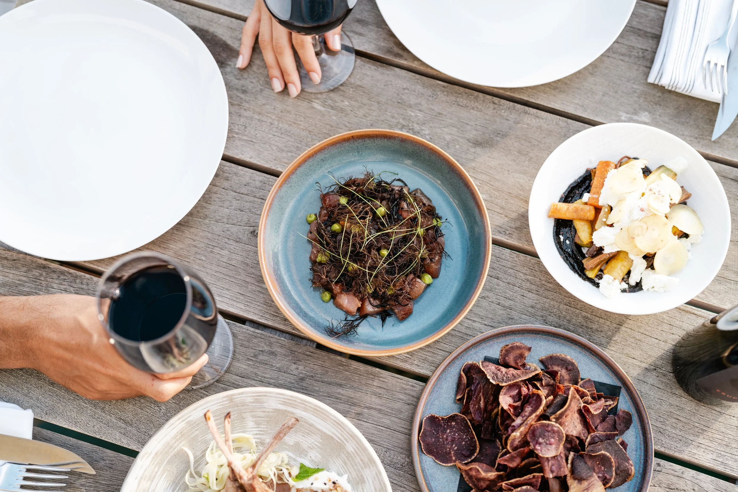 A top-down view of a wooden table with various plates of food and drinks. There are two glasses of red wine, a white plate, and three dishes with different types of food, including a plate of pasta with cauliflower and other vegetables, a salad with 