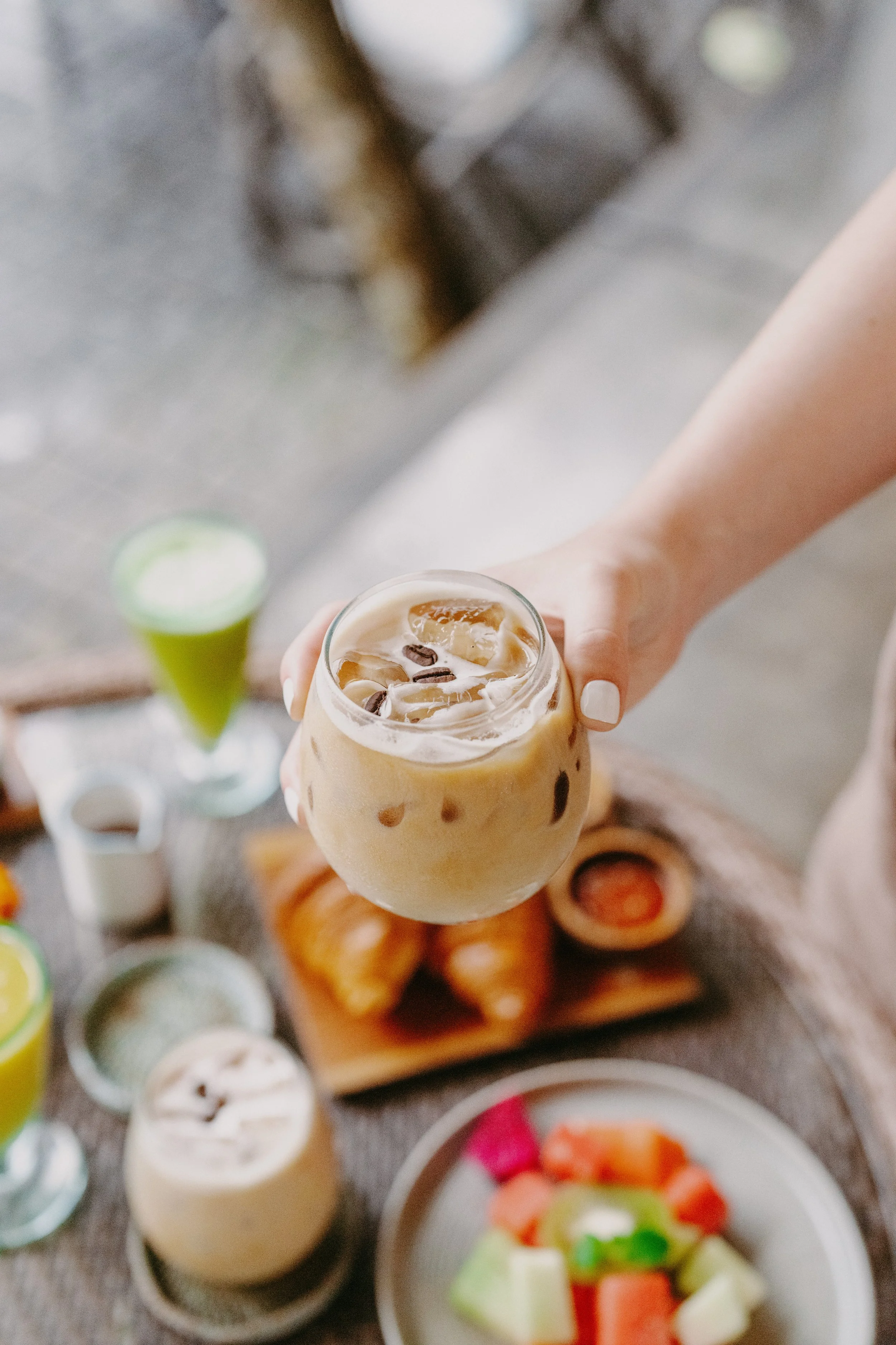 A person holding a glass of iced coffee with ice cubes and coffee beans on top, with a breakfast spread including croissants, fruit, and drinks on a table.